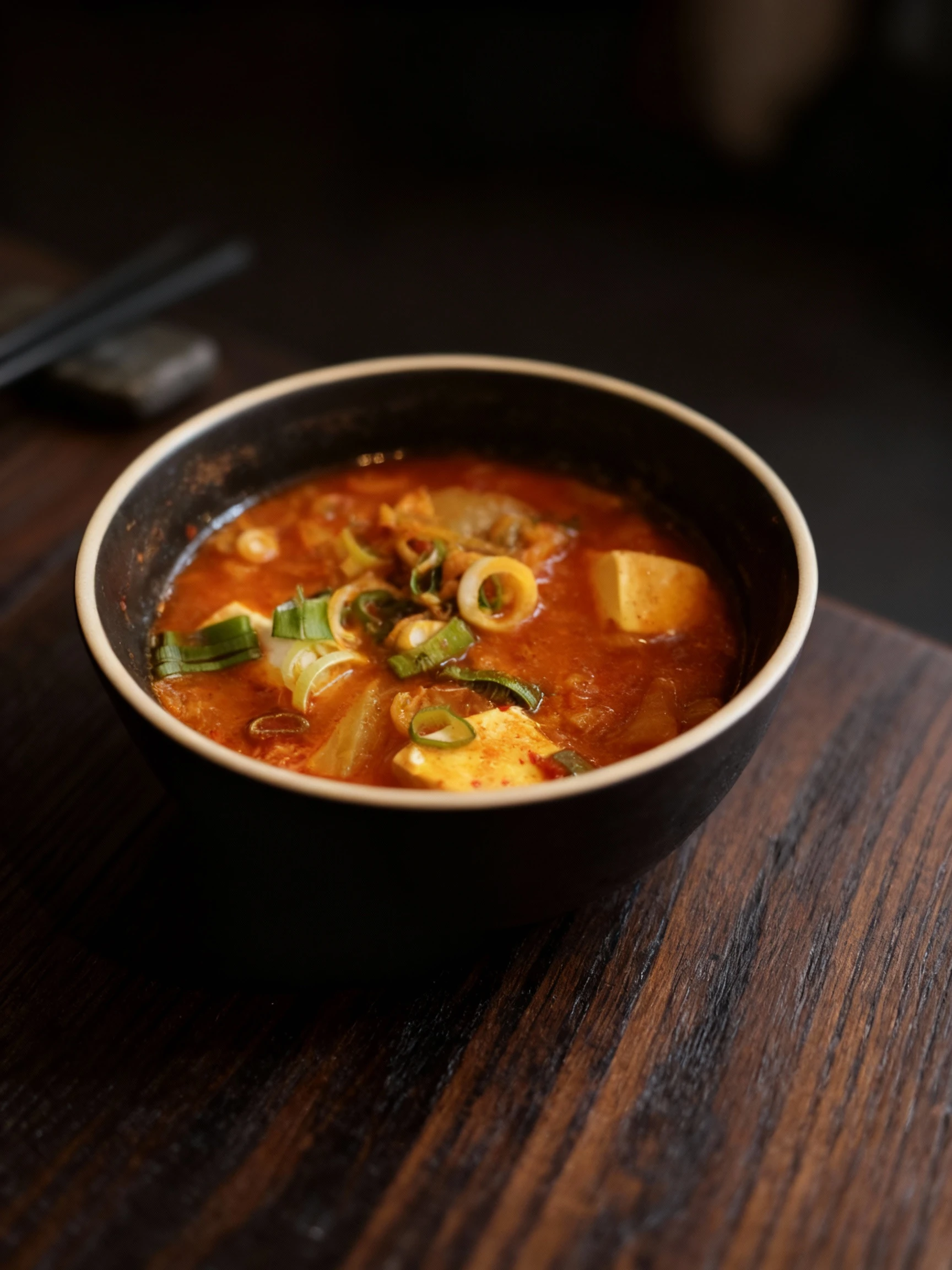 A bowl of Korean Kimchi-jjigae featuring soft tofu cubes, fermented kimchi, and sliced green onions in a spicy red broth.