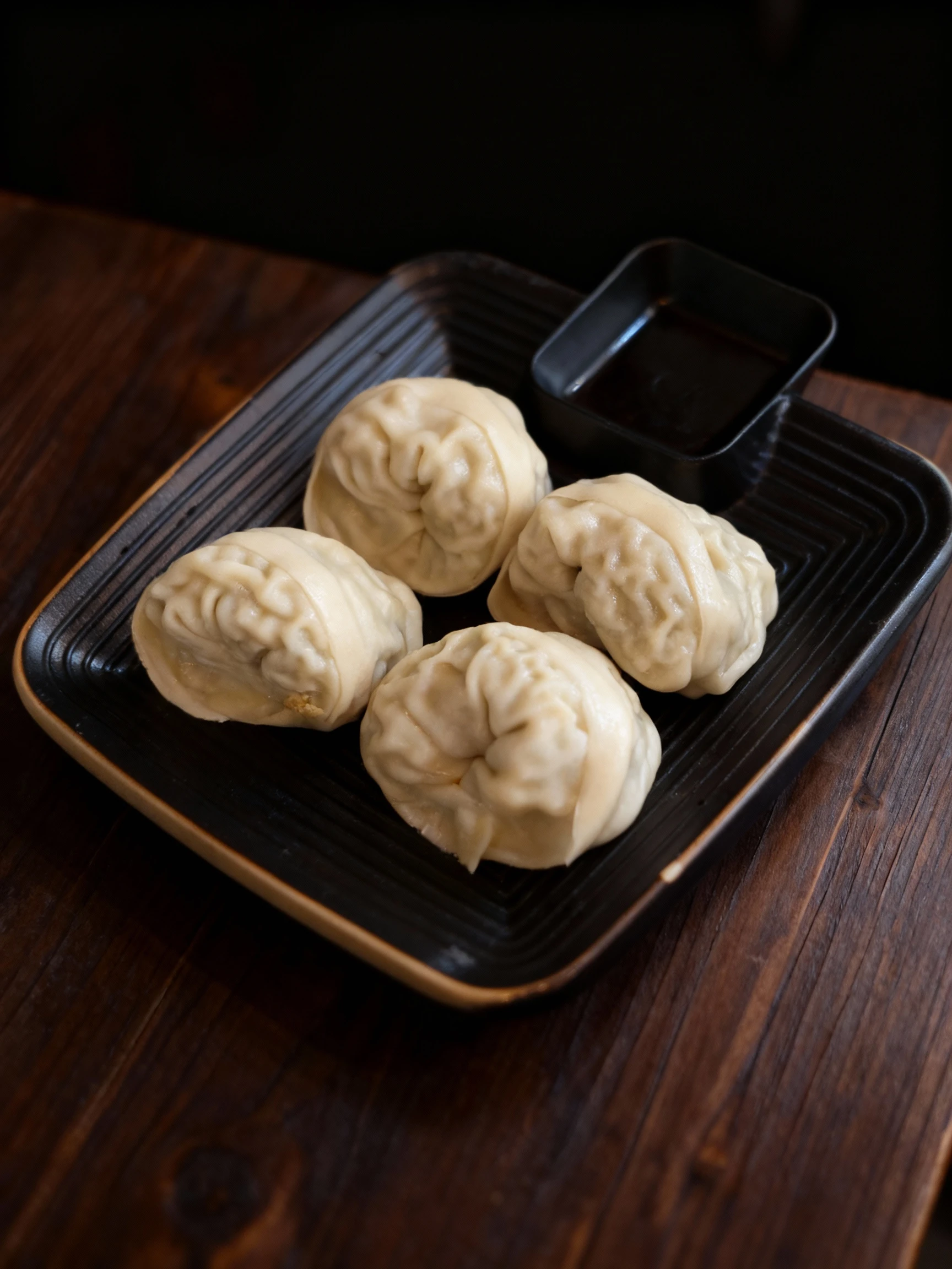 Four large steamed dumplings with thick, pleated dough served on a square black tray with an integrated sauce compartment.