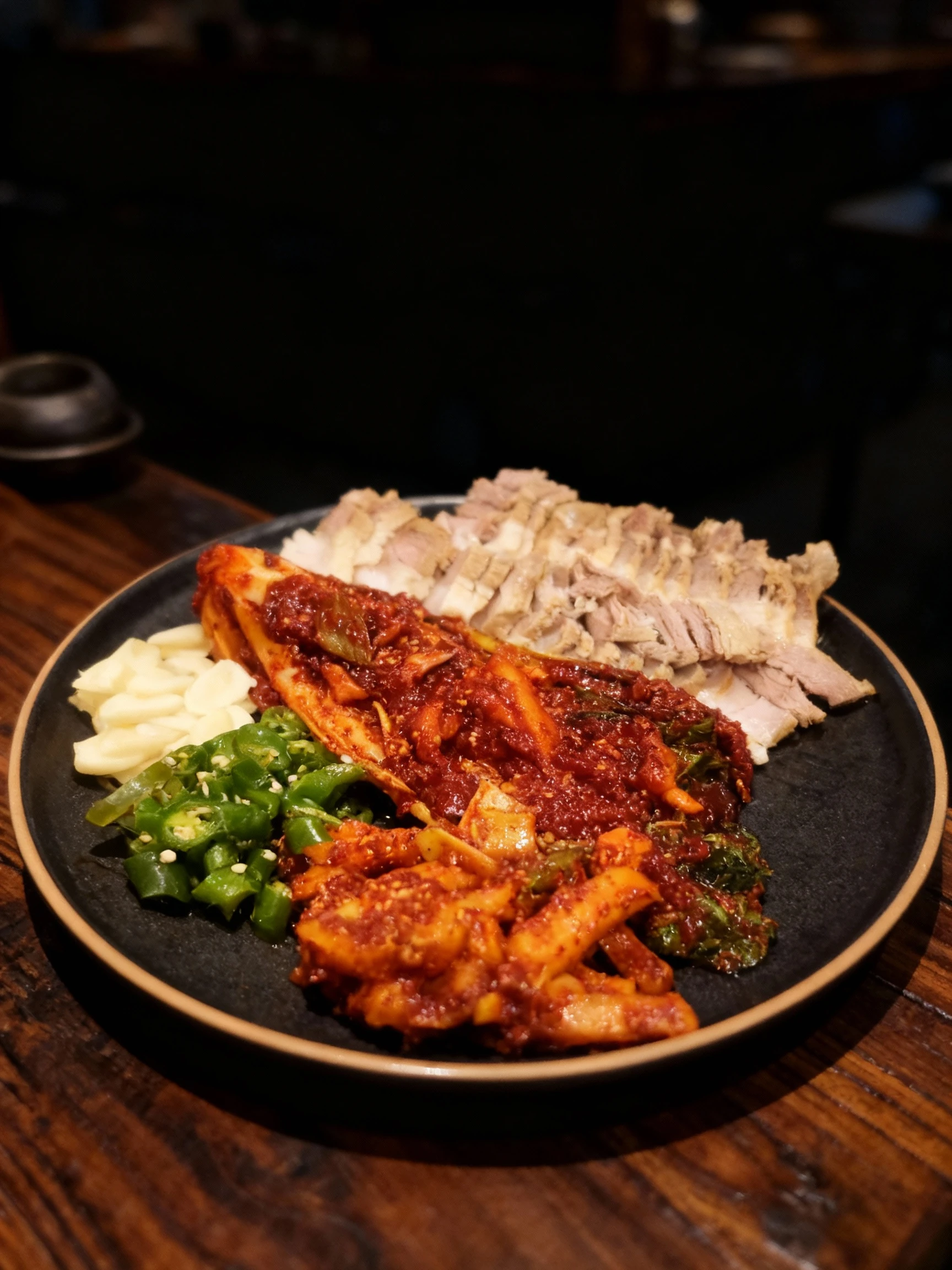 Bossam platter featuring sliced boiled pork belly served with spicy napa cabbage kimchi, seasoned radish, sliced raw garlic, and green chili peppers.
