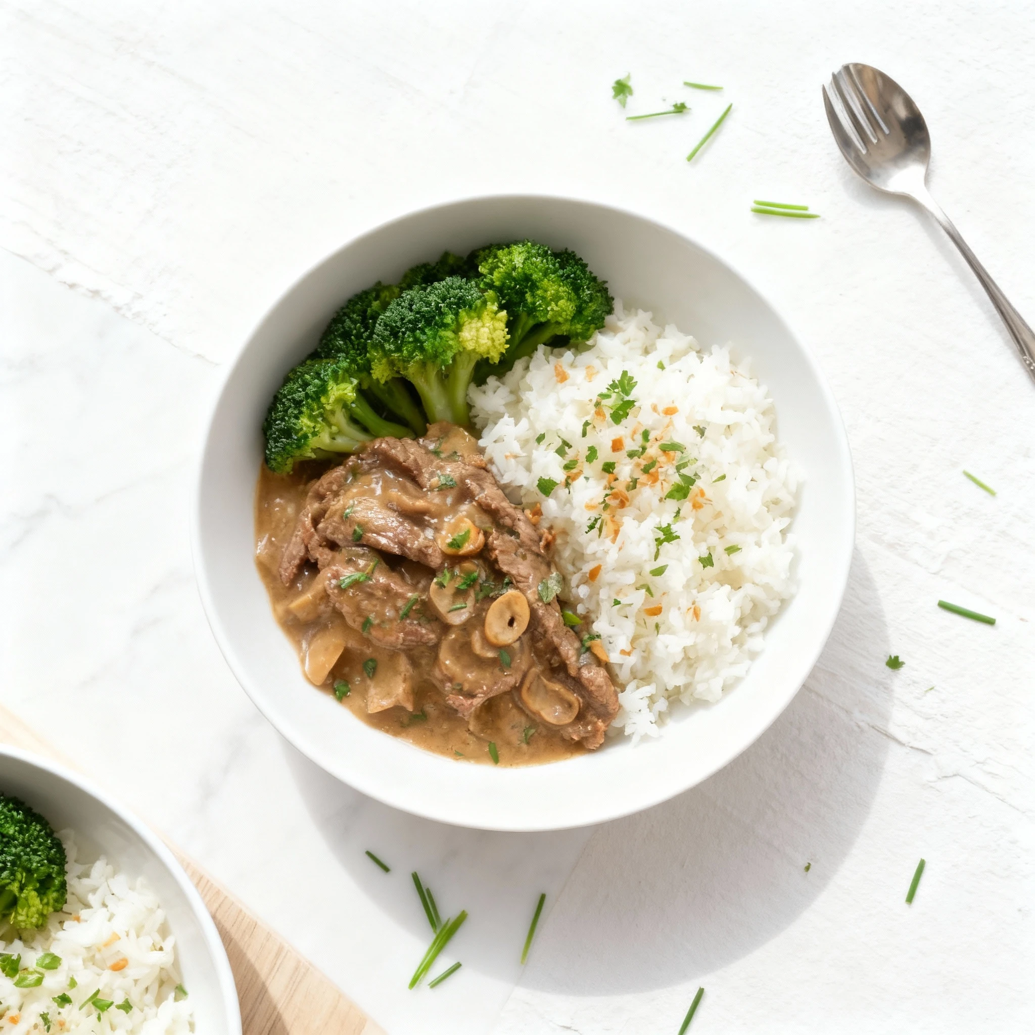Sliced beef and mushrooms in a creamy sauce are plated alongside a generous portion of white rice garnished with toasted garlic and herbs, and two florets of st