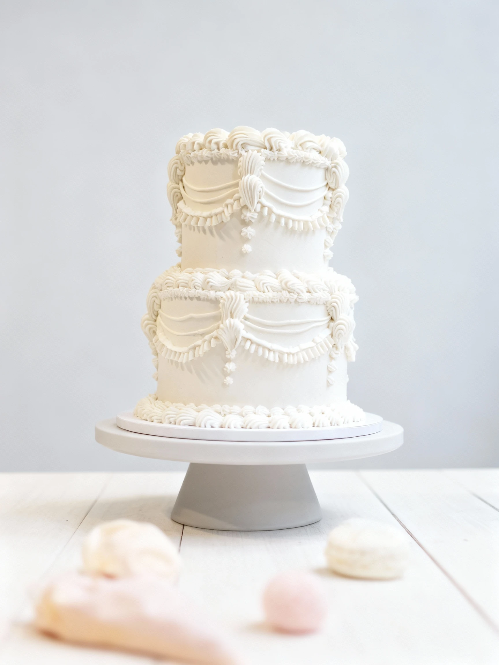 A classic two-tiered celebration cake covered in pale buttercream, featuring intricate, vintage-style Lambeth piping with detailed swags, ruffles, and shell bor