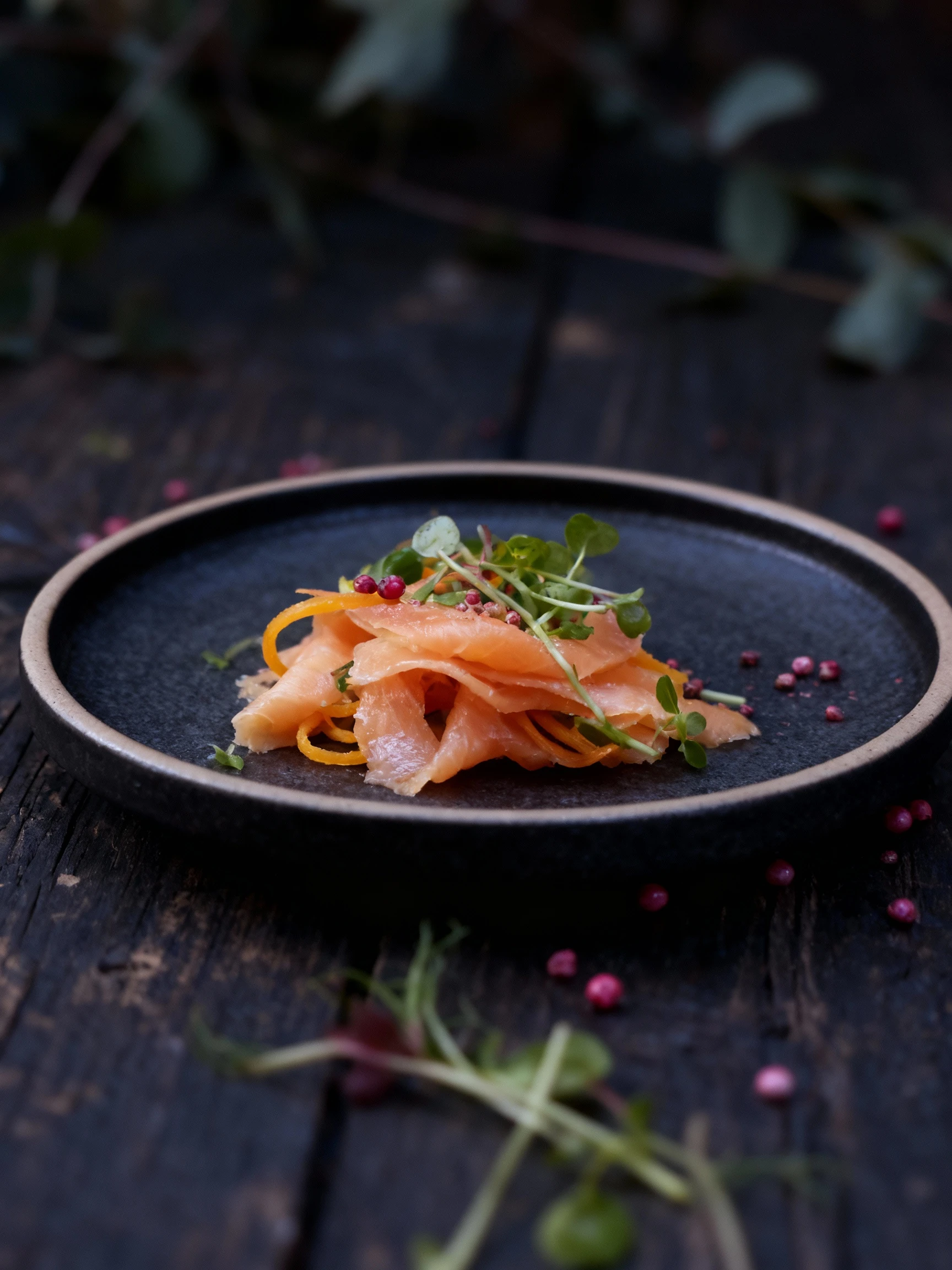 A starter dish of thinly sliced cured fish, likely salmon, topped with a tangle of spiralized carrots and a generous mound of mixed microgreens. The pale plate