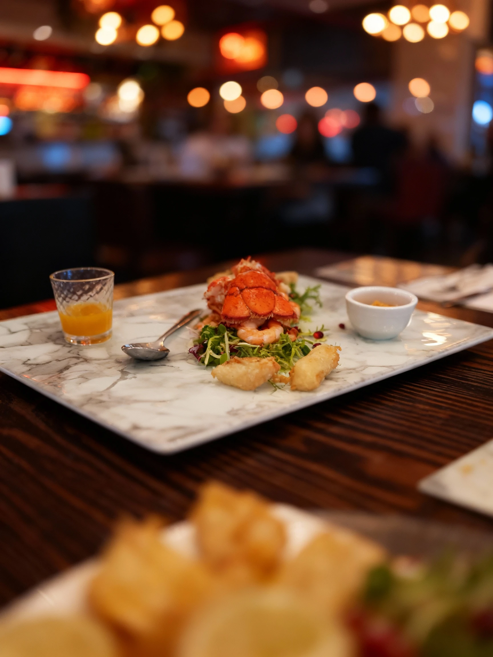 A seafood appetizer consisting of a lobster tail and peeled shrimp served atop a bed of mixed greens garnished with pomegranate seeds. The dish is accompanied b