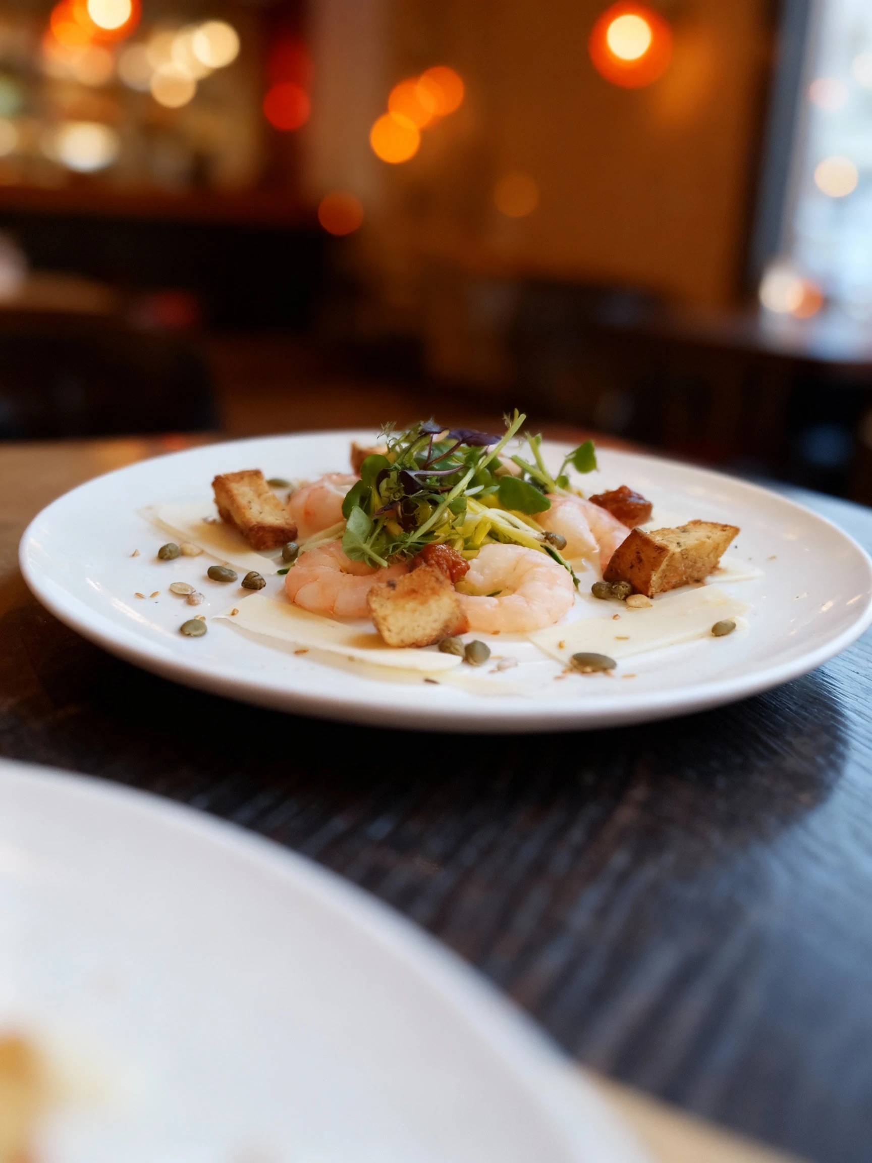 A gourmet appetizer featuring large peeled shrimp served over shavings of hard cheese, topped with a nest of microgreens and zucchini ribbons. The dish is garni