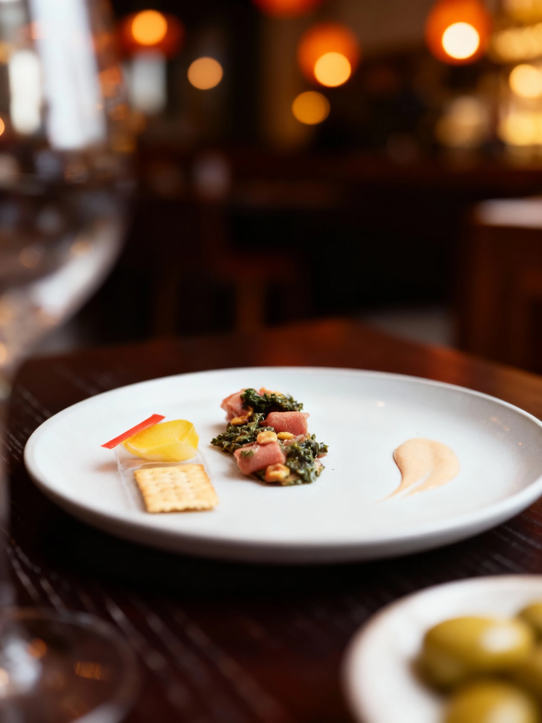 A composed appetizer featuring rolls of cured meat served over a dark green salad mixture, garnished with small yellow pieces, alongside a savory cracker and a