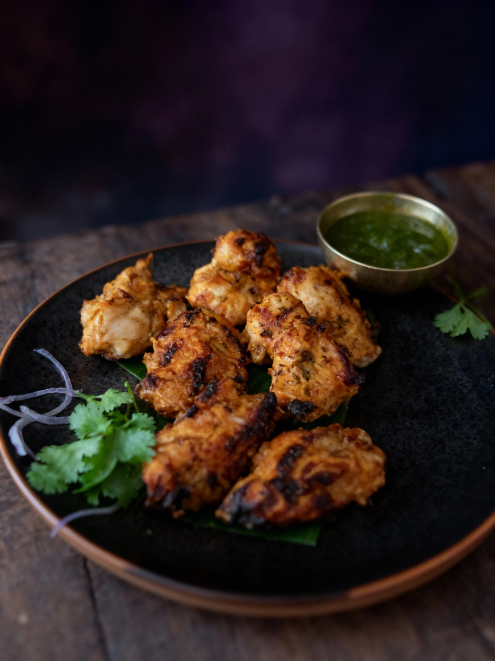 Grilled marinated chicken pieces, likely Chicken Tikka, served on a grey plate over a green leaf base, garnished with red onion slices, cilantro, and a lime wed