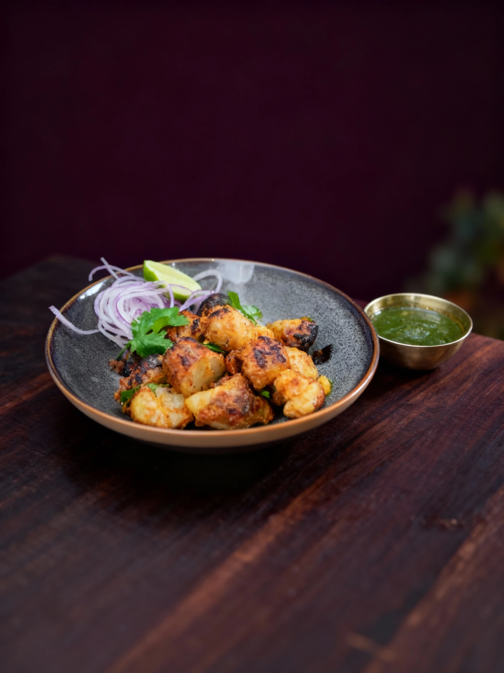 A serving of spiced and charred roasted vegetable pieces, resembling Gobi or Aloo Tikka, garnished with shredded red onion, fresh cilantro, and a lime wedge. Th