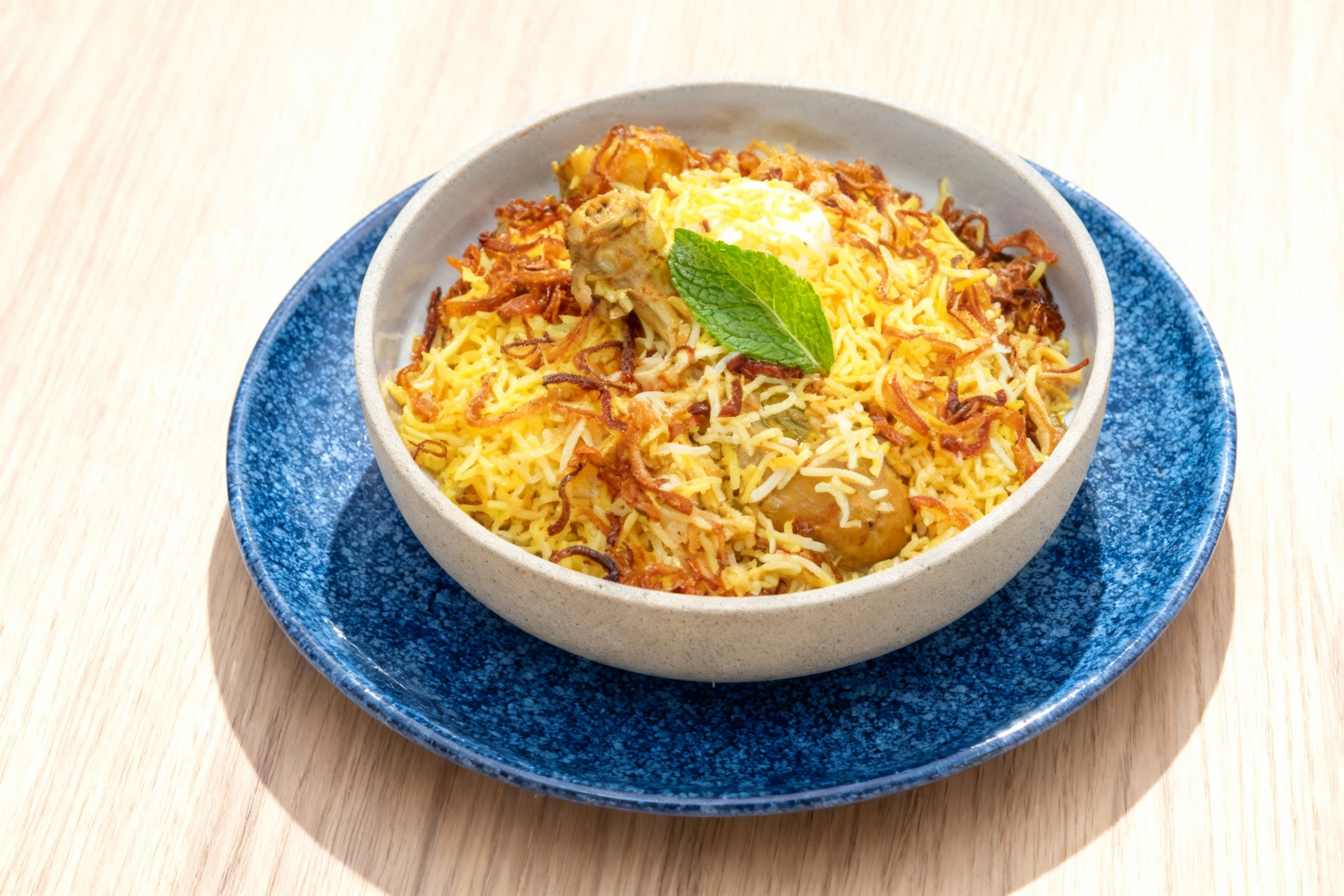 A serving of chicken Biryani featuring seasoned rice, pieces of chicken (including a visible drumstick), topped with crispy fried onions, a dollop of yogurt or