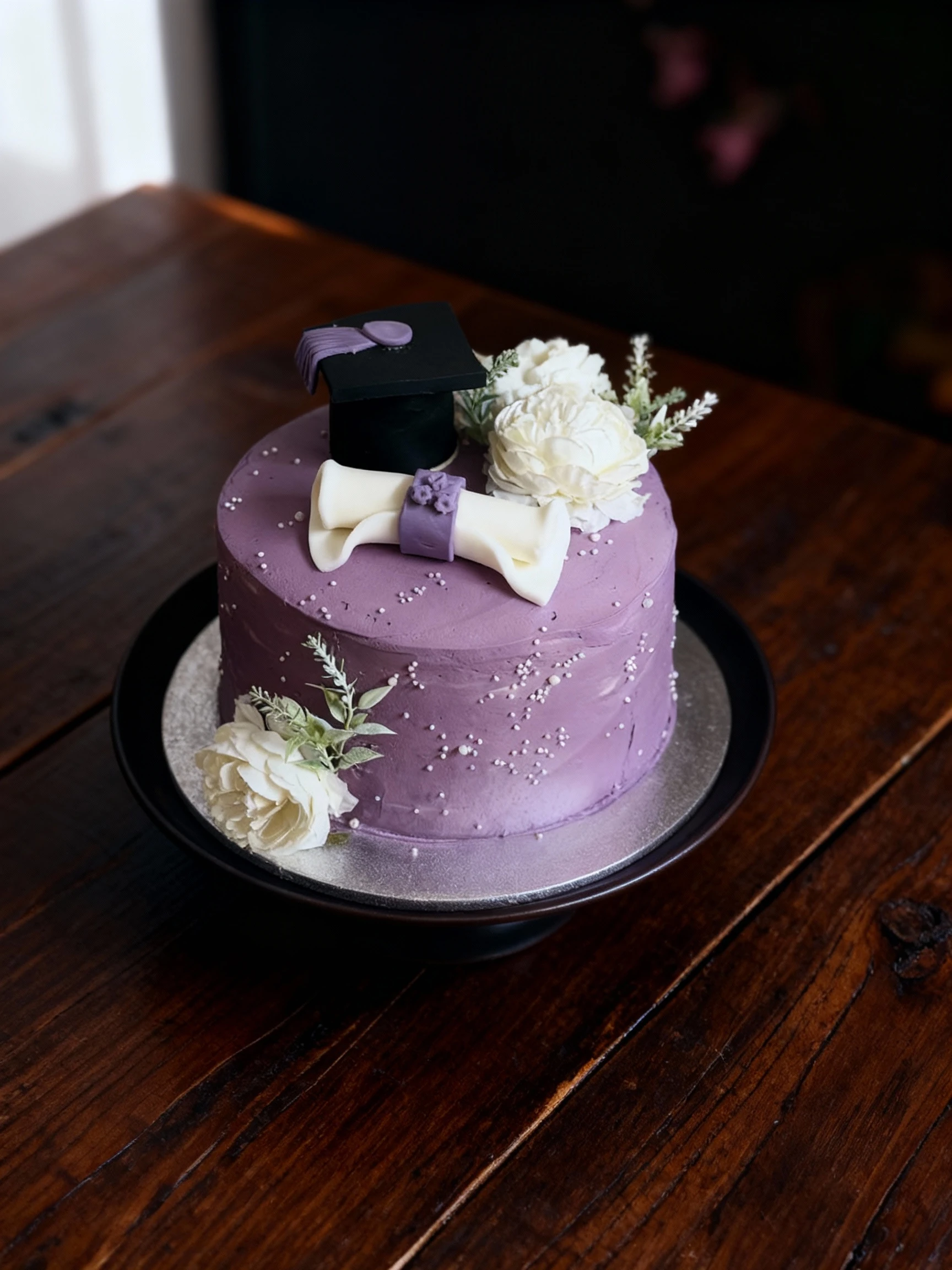 A round layer cake covered in textured lavender buttercream and white sprinkles, featuring graduation-themed fondant decorations including a black cap and a rol