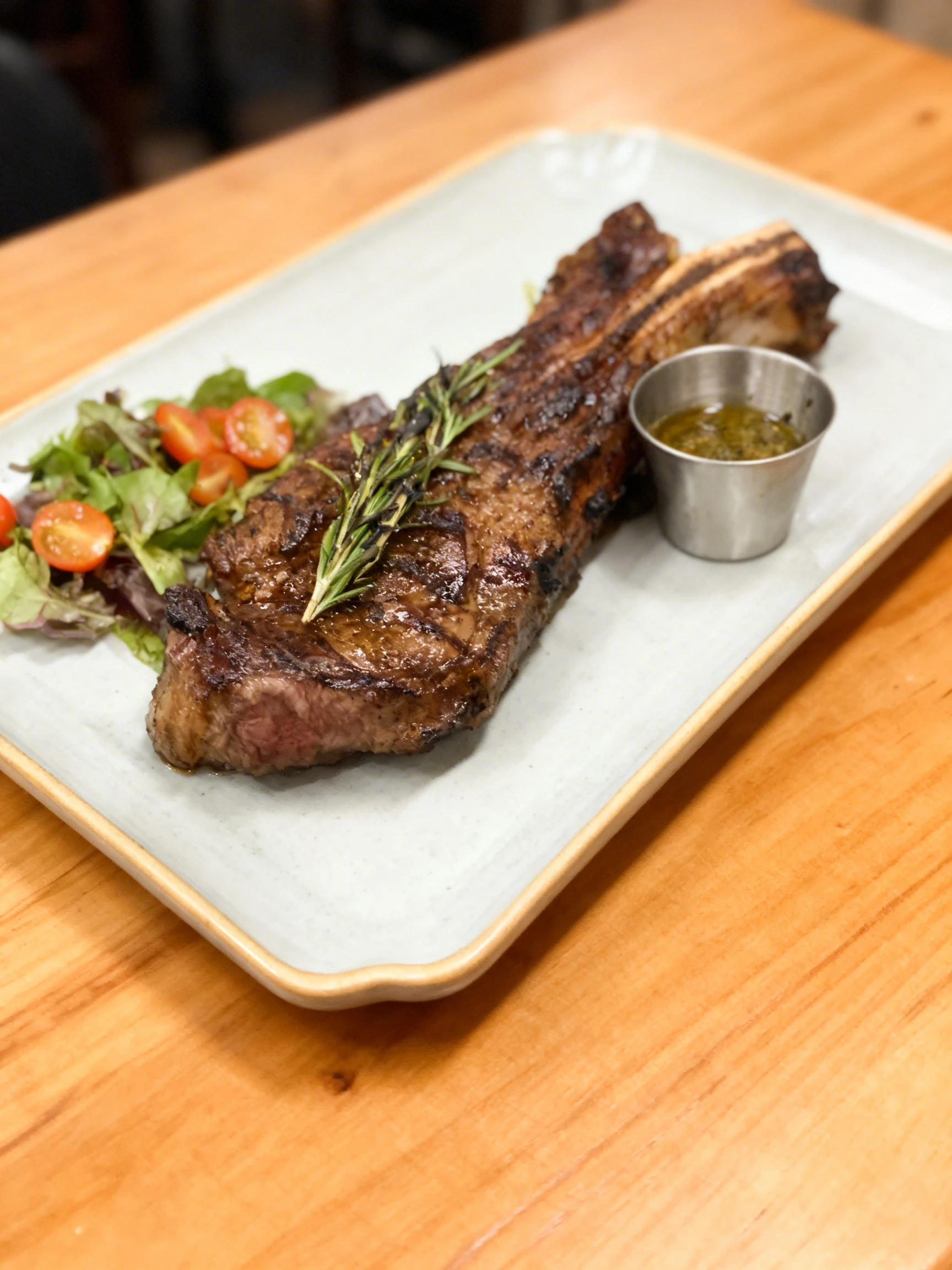 Grilled bone-in ribeye steak garnished with a charred rosemary sprig, served with a side of chimichurri and a garden salad with cherry tomatoes.