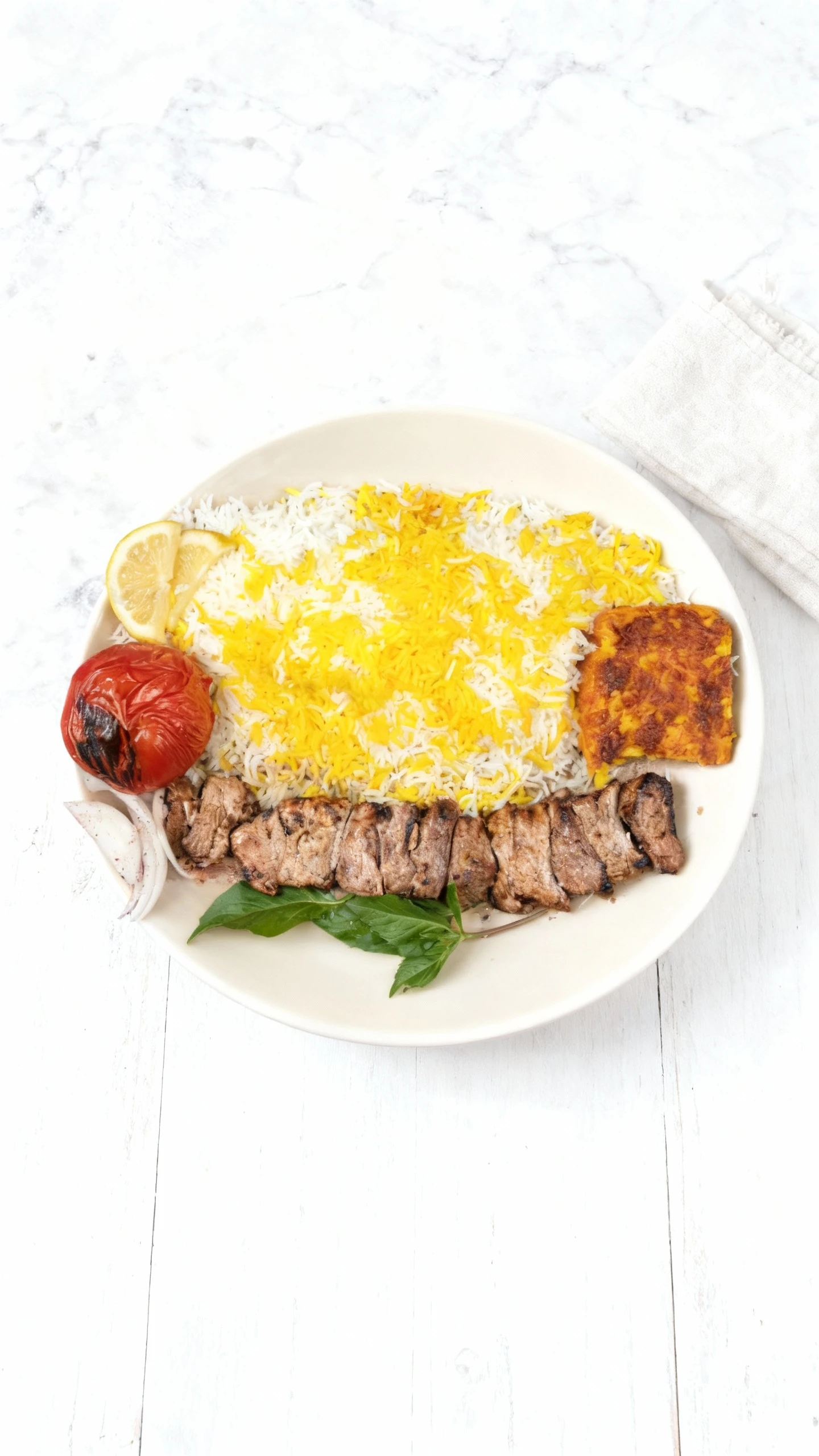 A plate of grilled kebab meat served alongside saffron-topped basmati rice, a rectangular piece of tahdig (crispy rice), a charred tomato half, lemon wedges, an