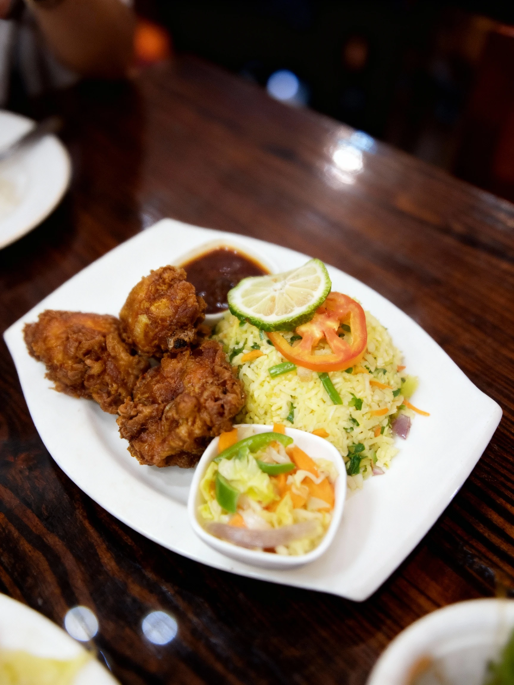 Crispy fried chicken pieces served with vegetable fried rice, a lime wedge, tomato slices, a side salad of cabbage and peppers, and a small dish of dark savory