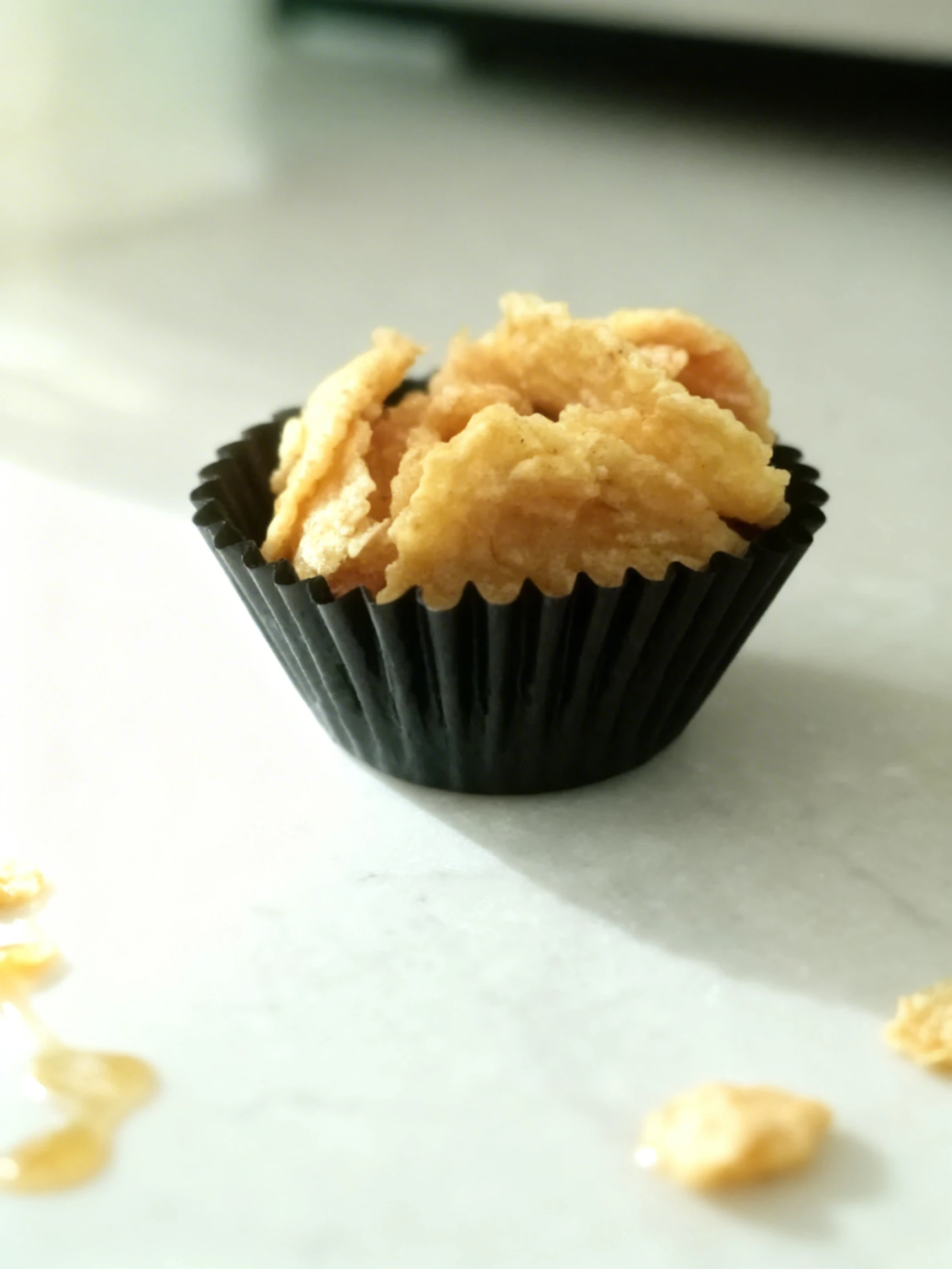 A honey cornflake cookie presented in a black pleated paper cupcake liner.