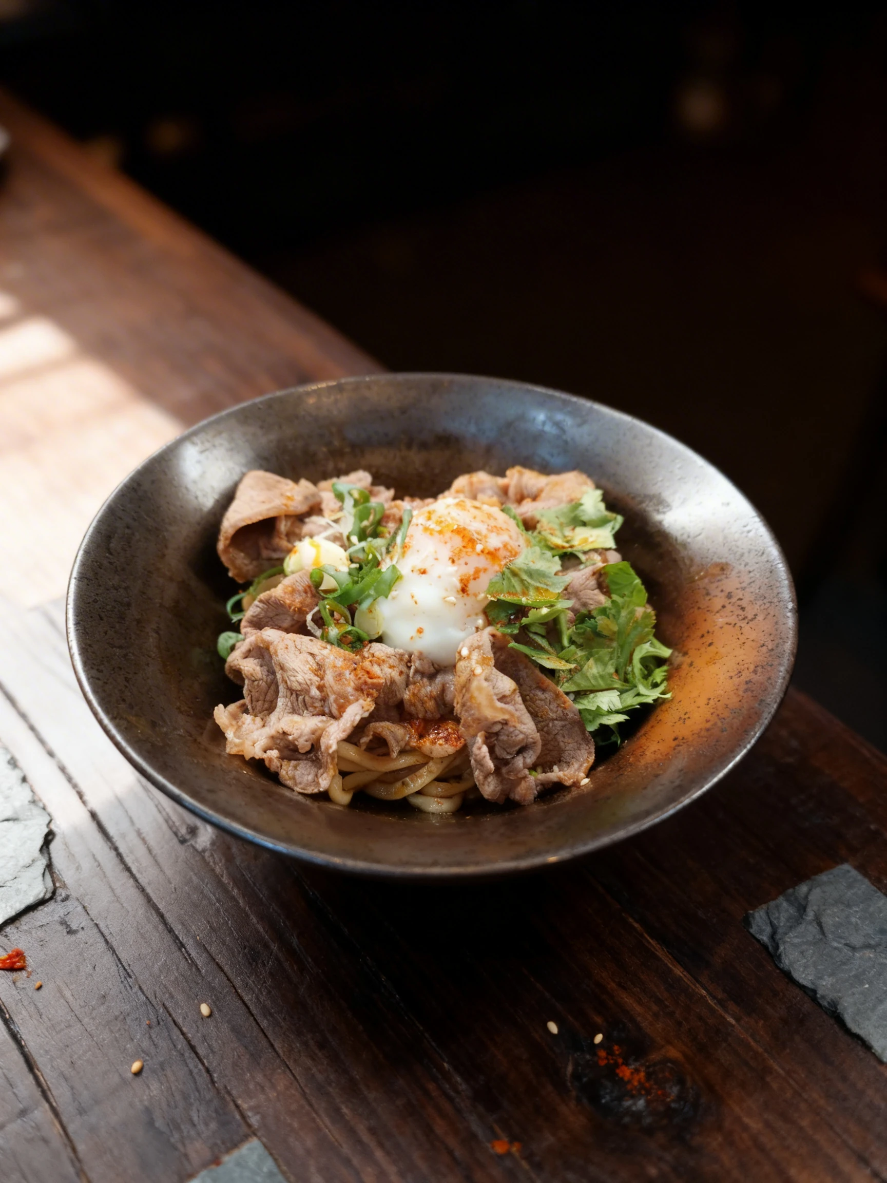 A beef bowl featuring thin slices of cooked beef over noodles, topped with a soft-boiled egg, fresh cilantro, scallions, sesame seeds, and chili powder.
