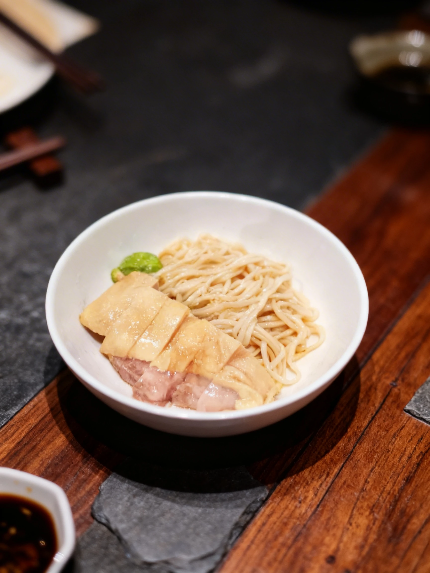 A bowl containing steamed chicken with skin sliced into thick pieces served alongside cooked wheat noodles and a side of green chili condiment.