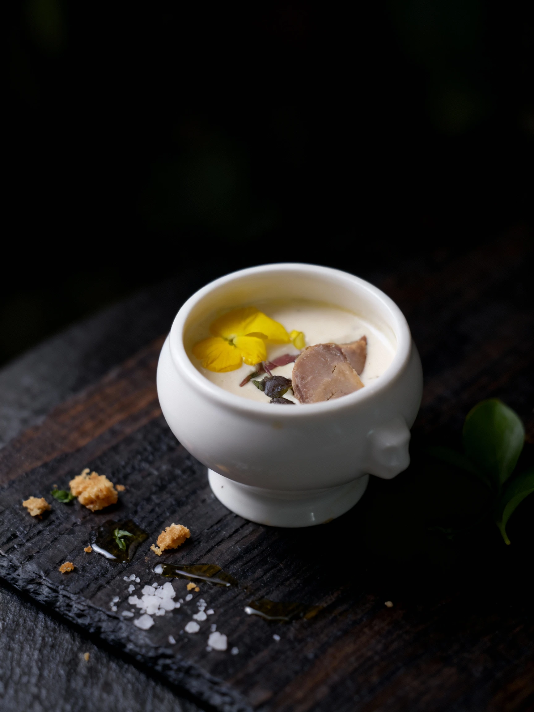 A creamy amuse-bouche or small portion of soup/velouté served in a white ceramic cup. The dish is garnished attractively with a slice of what appears to be pâté