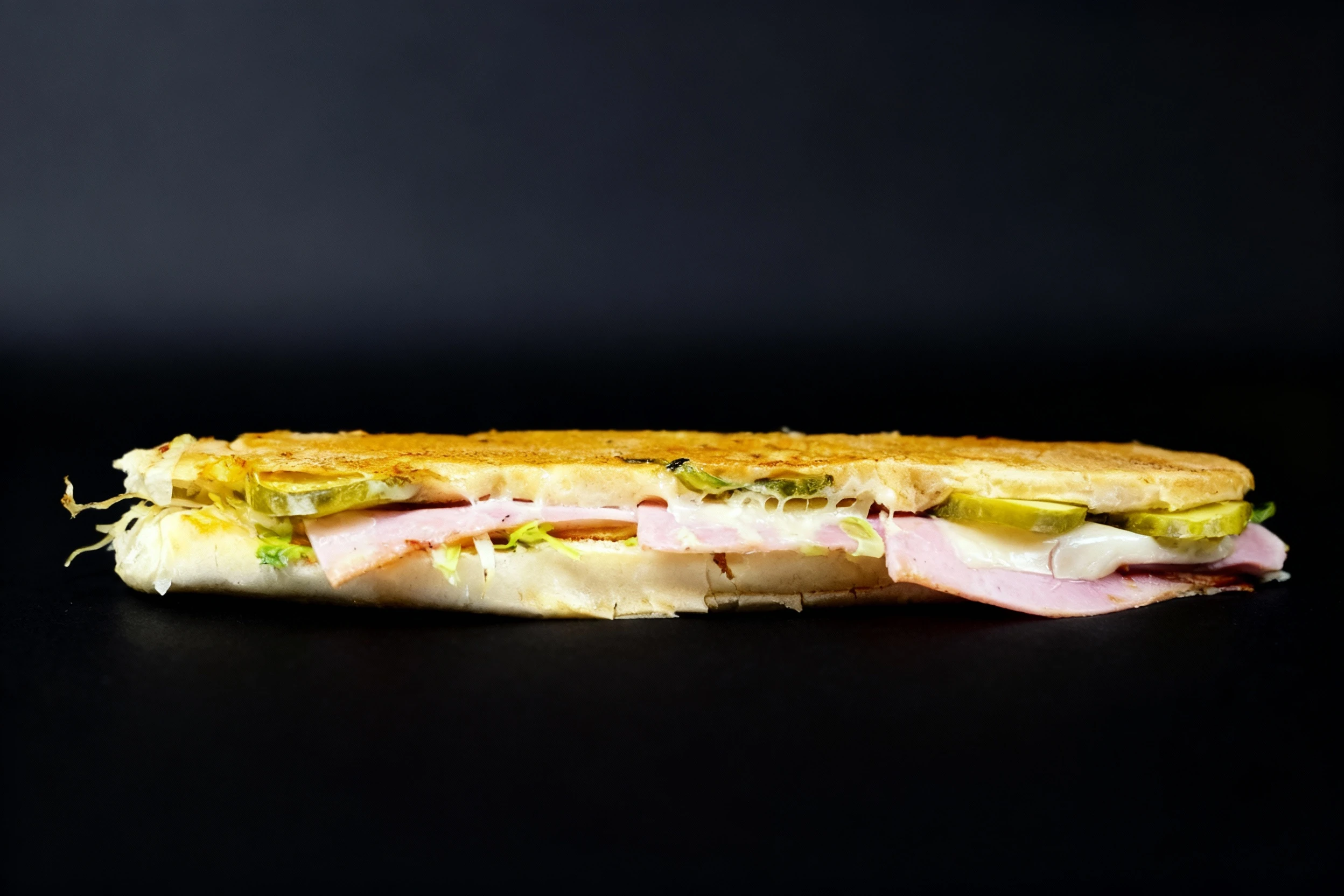 A long, pressed, and toasted sandwich, likely a Cuban or panini variation, filled with sliced ham, melted white cheese, dill pickles, and shredded lettuce.