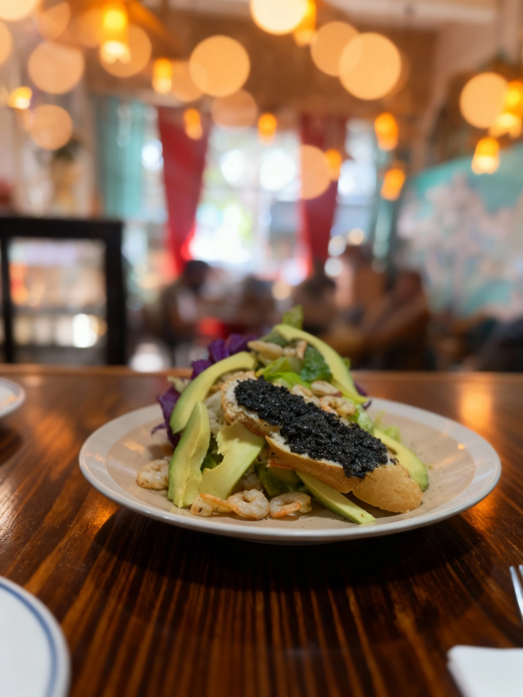 A seafood salad consisting of grilled shrimp, sliced avocado, and mixed greens, topped with a piece of toasted baguette spread with black olive tapenade.