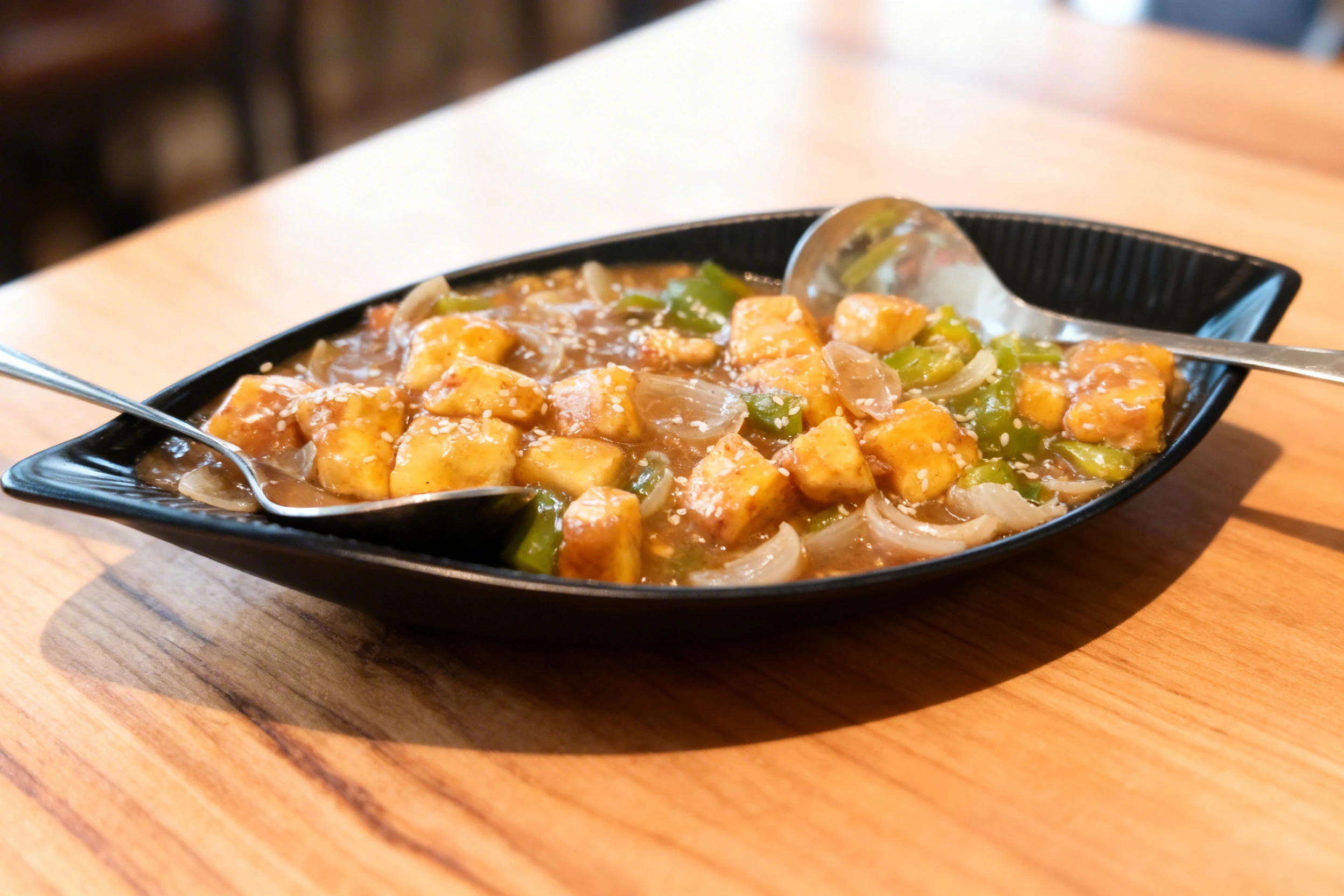 Chilli paneer cubes with sautéed green peppers and onions in a thick savory sauce, topped with white sesame seeds and served in a black oval dish.