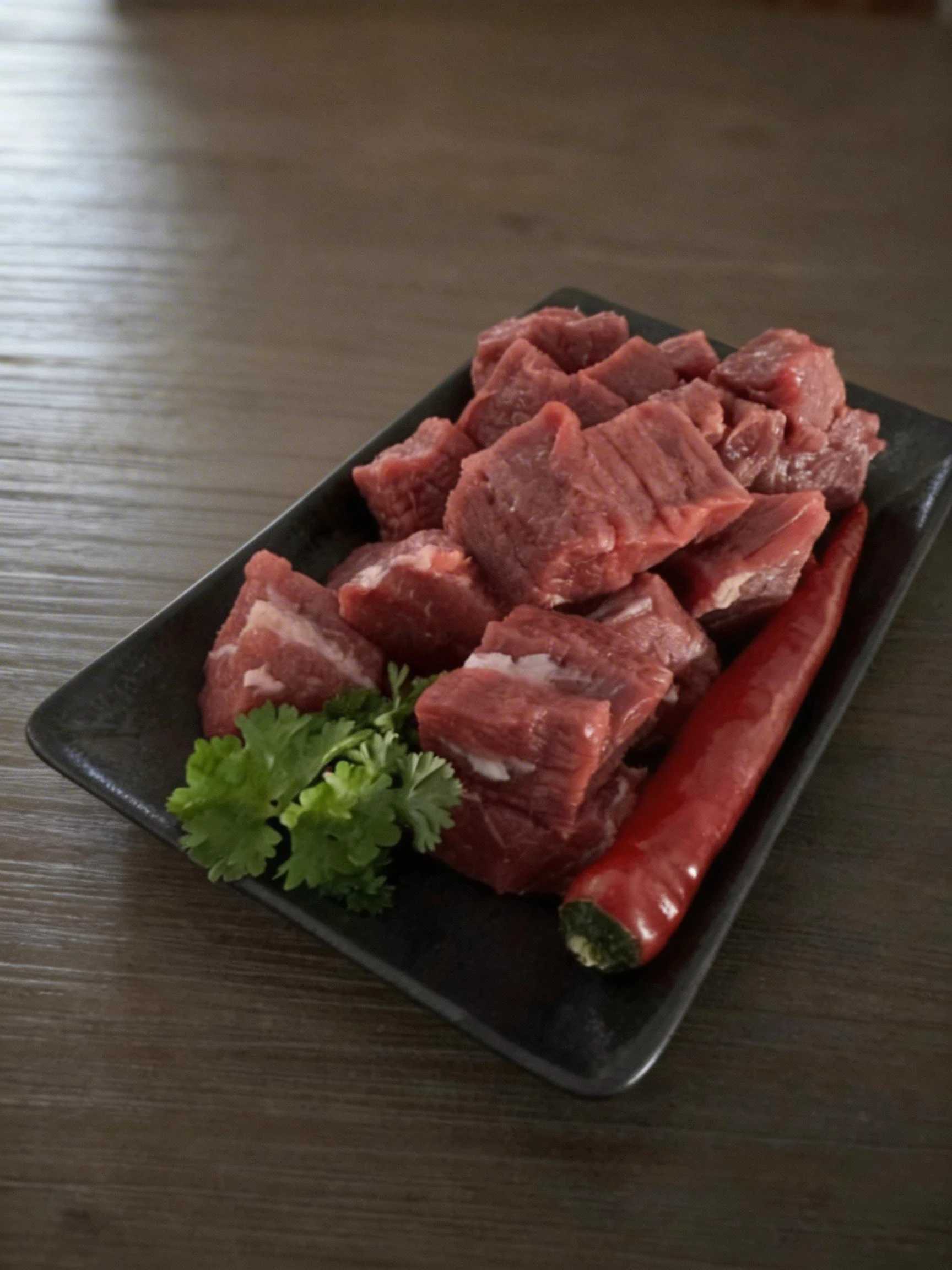 Cubed raw red meat, likely beef or lamb, served on a black tray with a whole red chili pepper and a sprig of fresh cilantro.