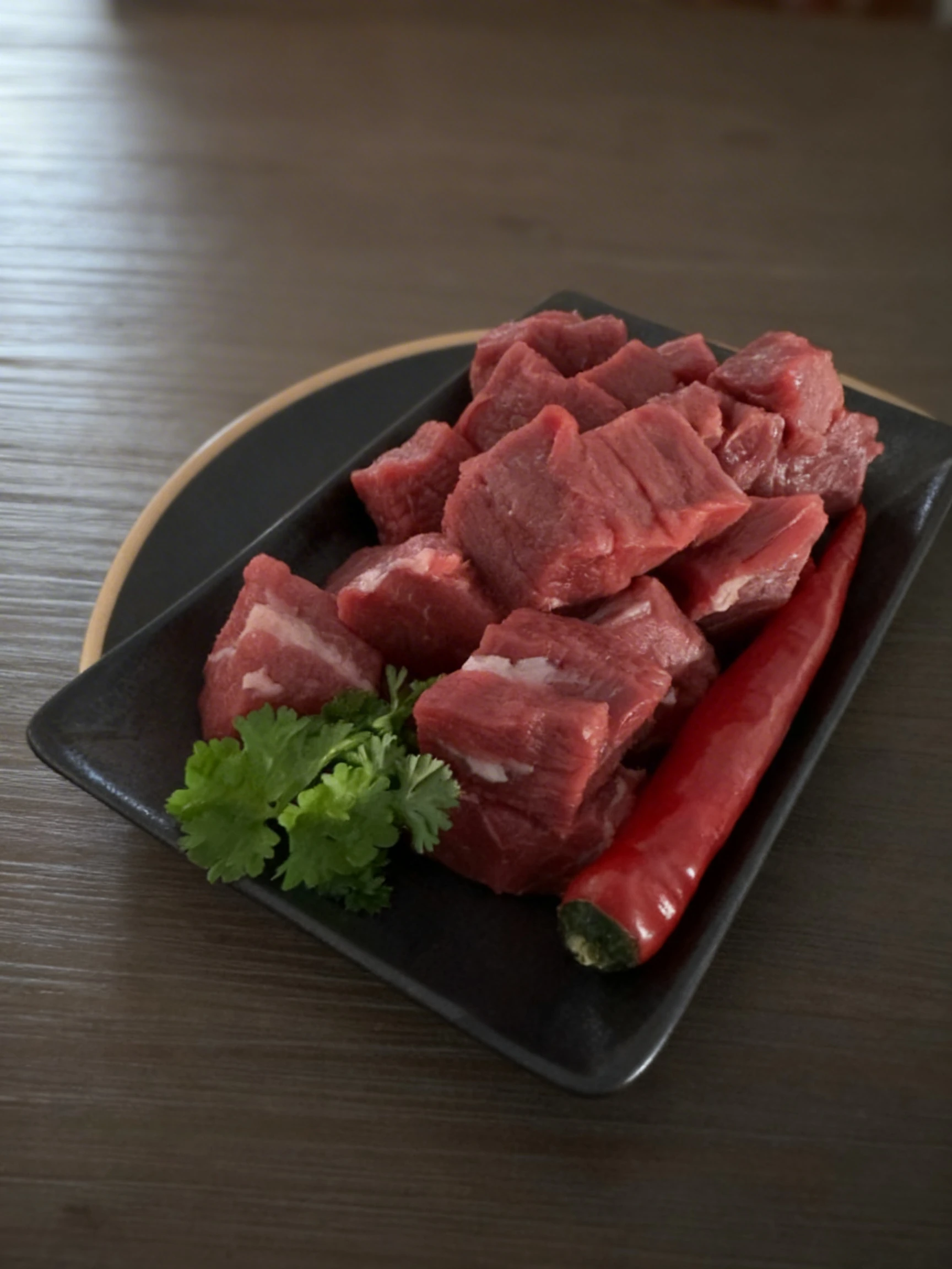 Cubed raw red meat, likely beef or lamb, served on a black tray with a whole red chili pepper and a sprig of fresh cilantro.
