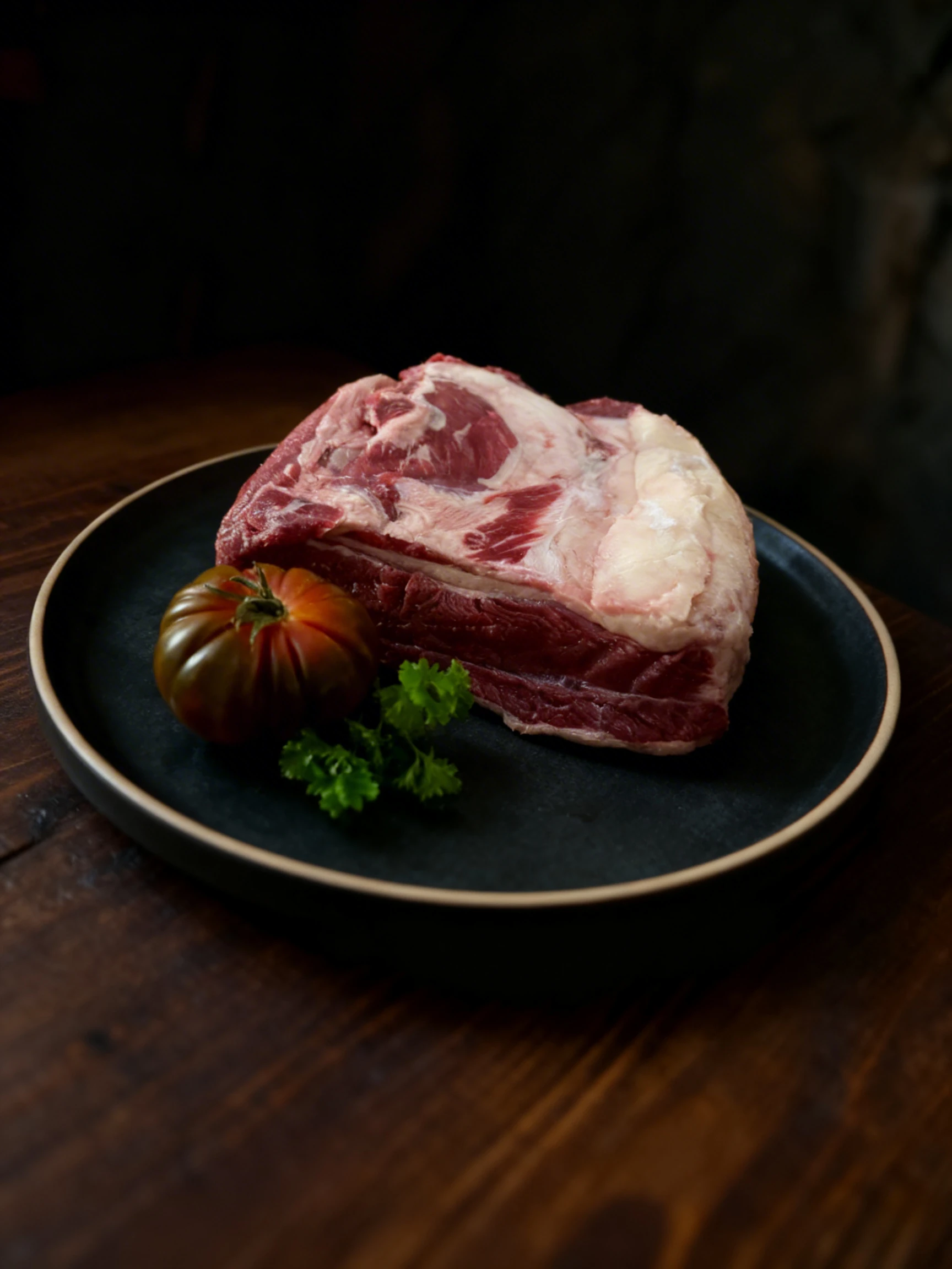A large cut of raw beef ribeye with significant fat marbling and a thick fat cap, accompanied by a whole heirloom tomato and a sprig of fresh parsley on a slate