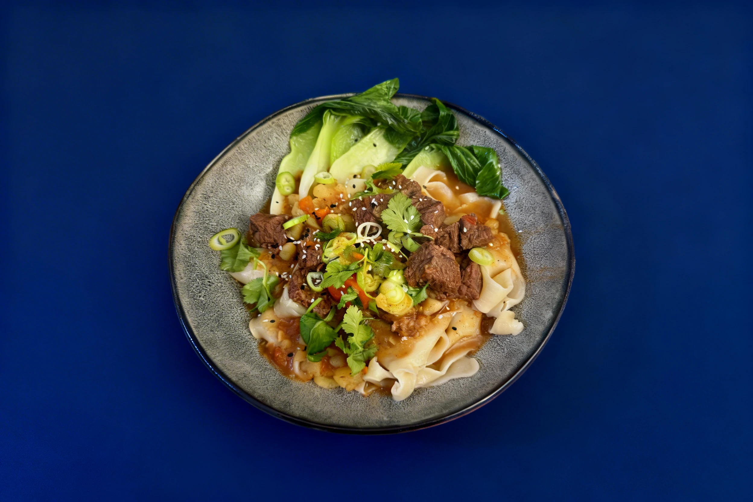 A bowl of wide rice noodles topped with braised beef chunks, bok choy, chopped scallions, fresh cilantro, and sesame seeds in a savory sauce.
