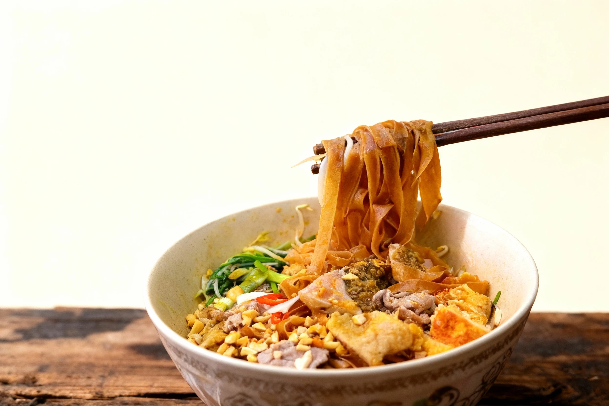A bowl of dry flat rice noodles featuring sliced meat, fried tofu pieces, bean sprouts, and green vegetables, heavily garnished with crushed peanuts and red chi