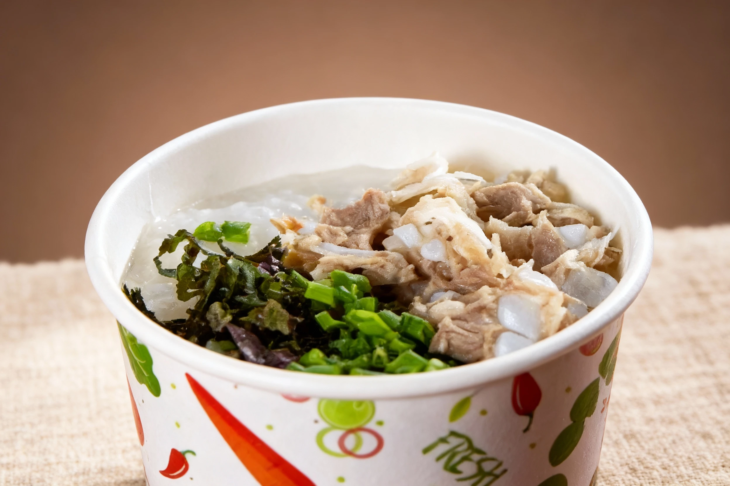 A savory meal, likely a Vietnamese-style rice porridge (cháo) or noodle soup component, served in a disposable cup and topped with shredded white meat, chopped