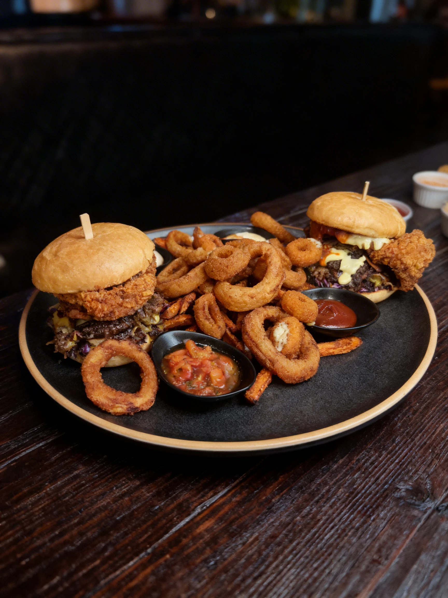 A large shared platter featuring two burgers with fried chicken and beef patties, accompanied by a mountain of onion rings, sweet potato fries, mozzarella stick