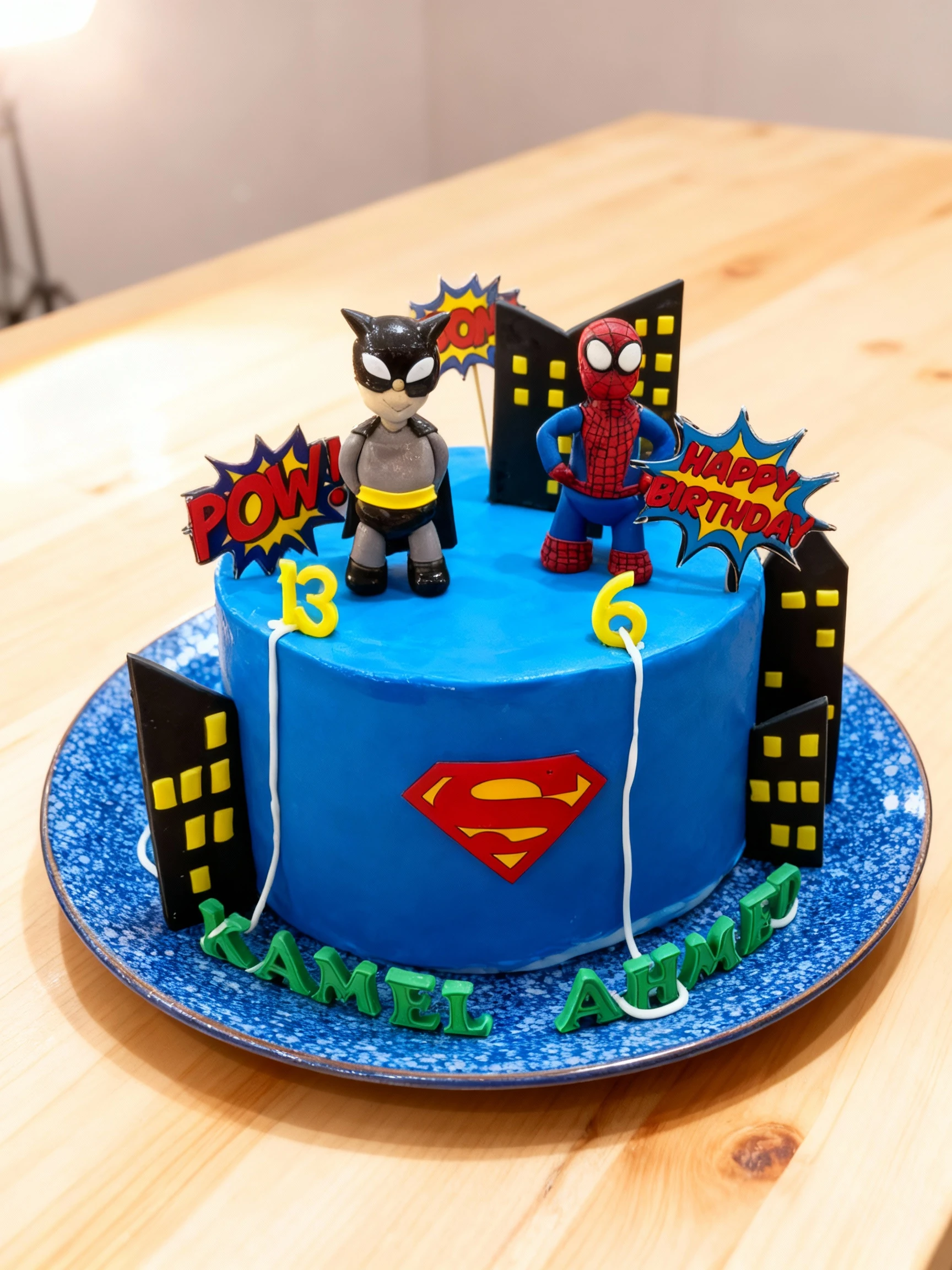 A single-tier superhero birthday cake covered in bright blue fondant, featuring a Superman "S" shield logo on the side and topped with Batman and Spiderman figu