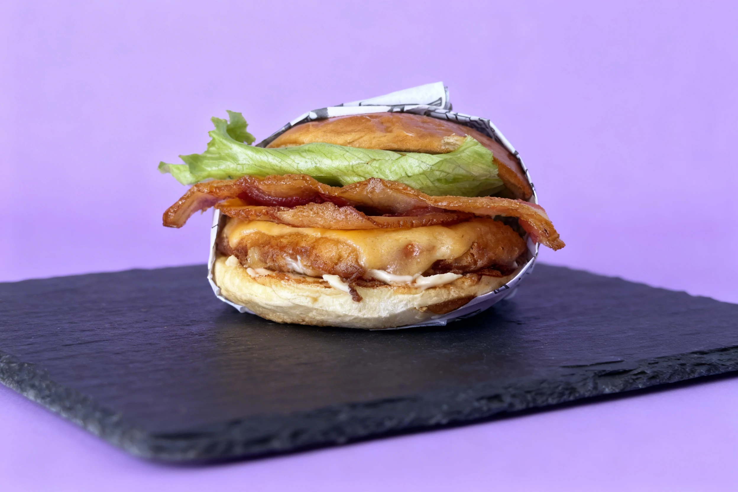 A chicken sandwich featuring a breaded chicken patty, melted American cheese, crispy bacon slices, and iceberg lettuce on a brioche bun.