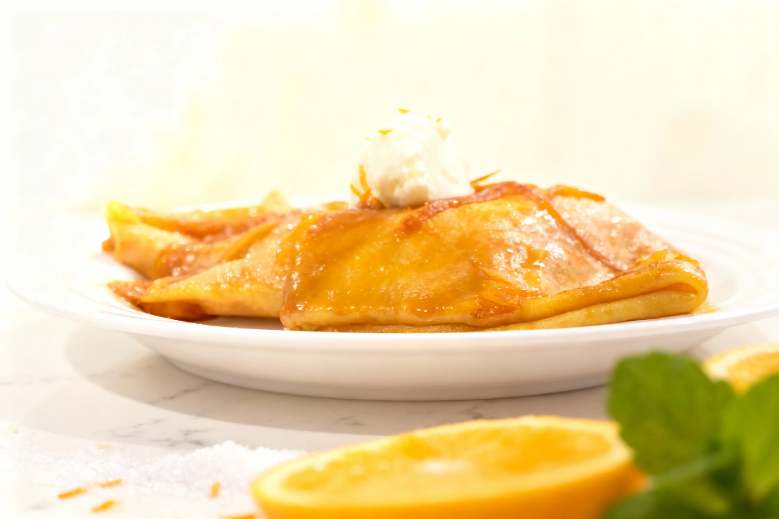 Folded dessert crepes coated in a thick, glossy orange or caramel glaze, featuring visible strands of citrus zest, and topped with a dollop of white cream or ic
