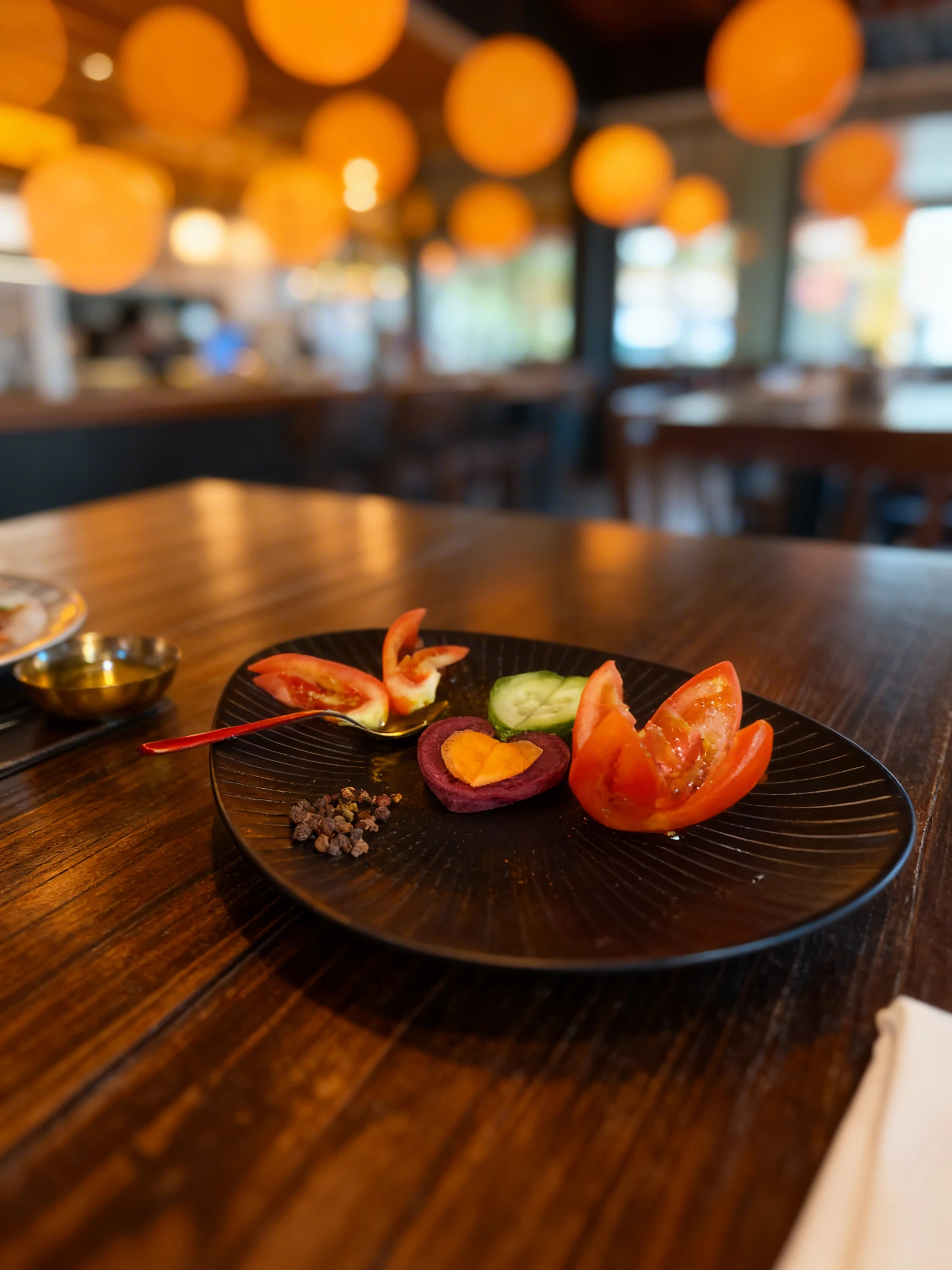 Artistic arrangement of sliced tomatoes, cucumbers, and heart-shaped beets on a black textured plate, accompanied by a small gold bowl of oil and scattered whol