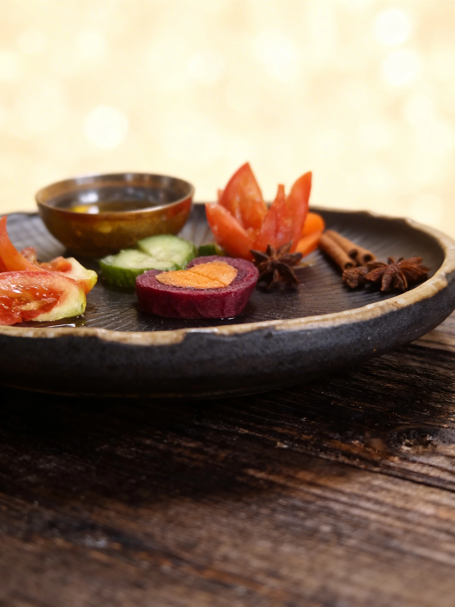 Sliced vegetable garnish featuring tomato wedges, cucumber slices, and heart-shaped beetroot with carrot, accompanied by a bowl of oil and scattered whole spice