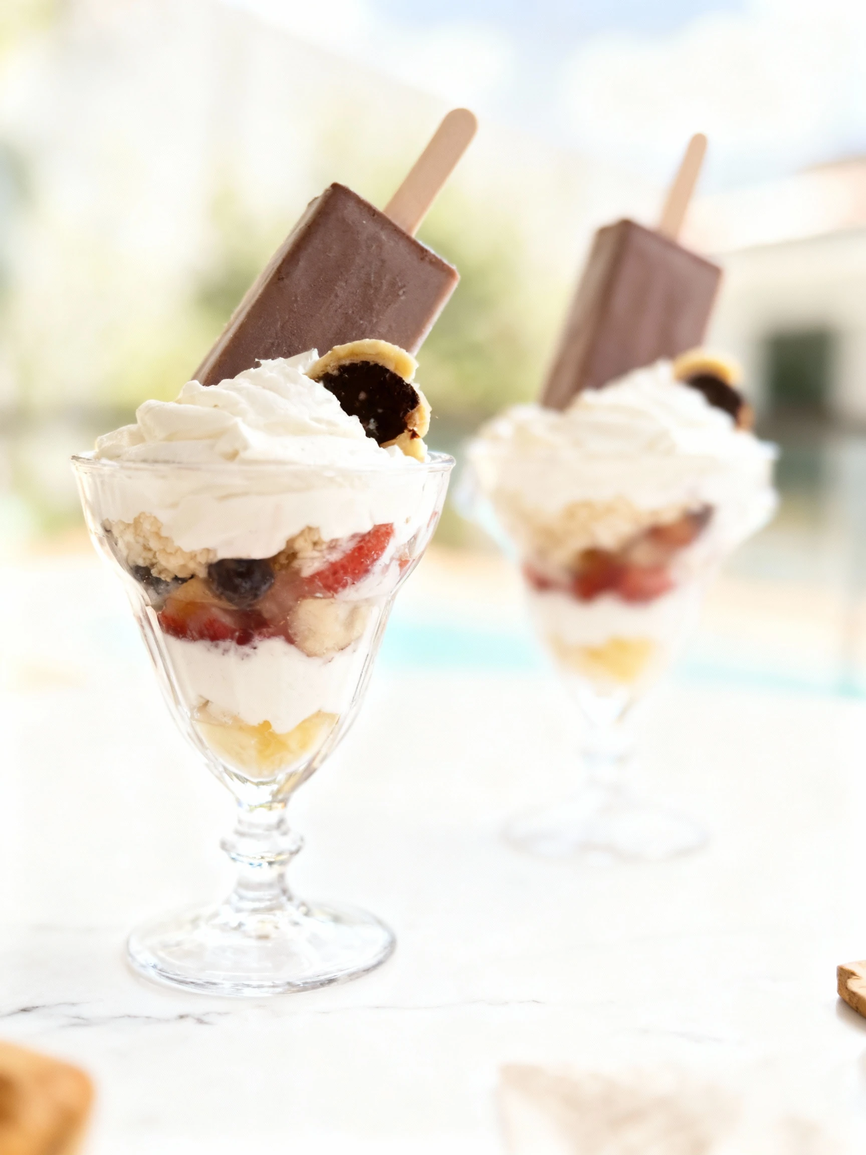 Gourmet dessert sundaes in plastic cups featuring layers of cream, fruit, and chocolate, topped with whipped cream and whole frozen ice cream bars on sticks.