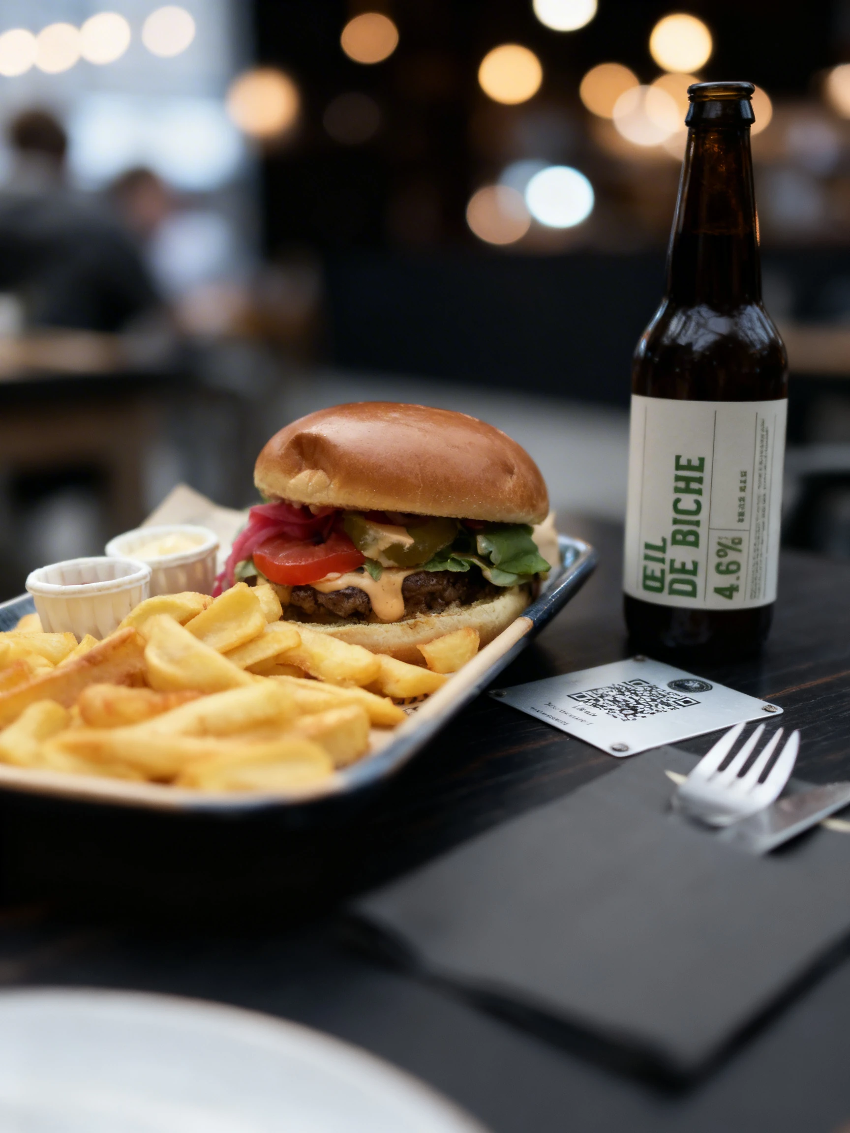 Beef burger on a brioche bun with tomato, pickles, pickled red onions, and sauce, served with thick-cut fries and side containers of ketchup and mayonnaise.