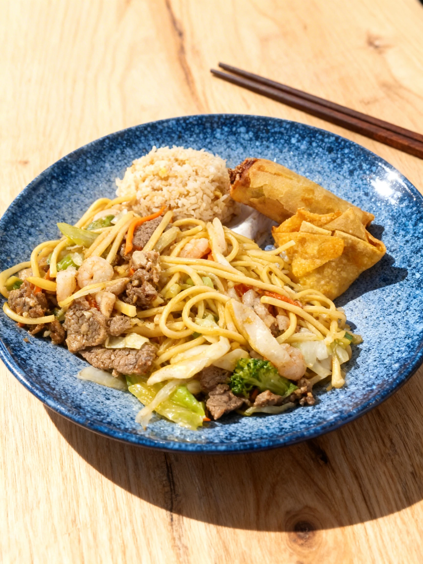 A combination platter of lo mein with beef, shrimp, and vegetables, served with a side of fried rice, a spring roll, and fried wontons.