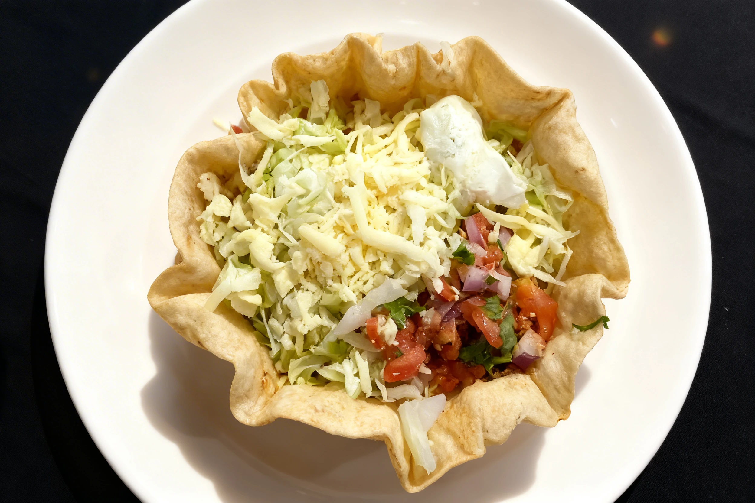 A taco salad served in a fried flour tortilla bowl, containing shredded lettuce, diced tomatoes, onions, cilantro, and topped with shredded cheese and sour crea