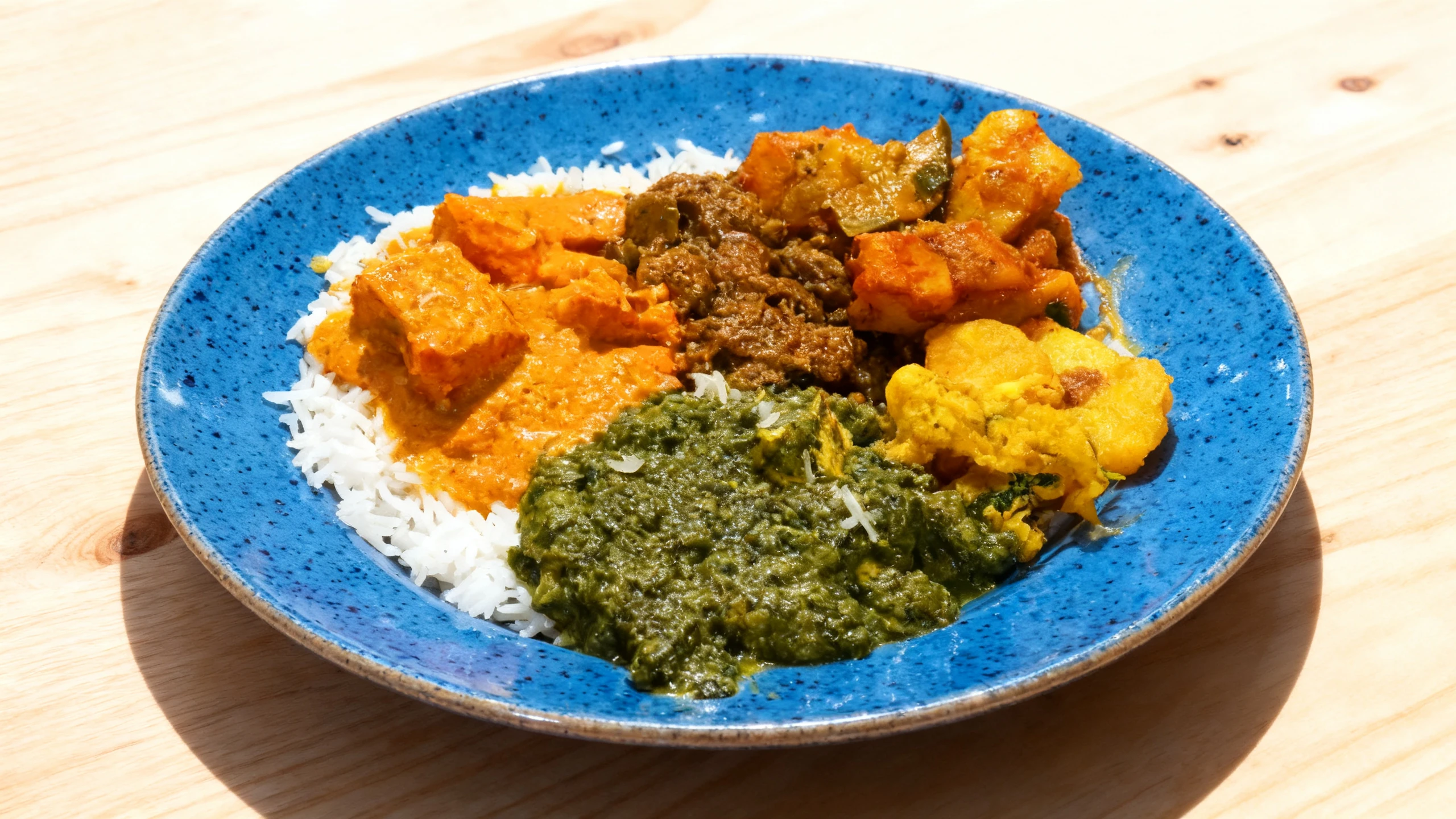A plate of Indian cuisine featuring saag paneer, butter chicken, and various vegetable curries served over white rice with fried fritters.