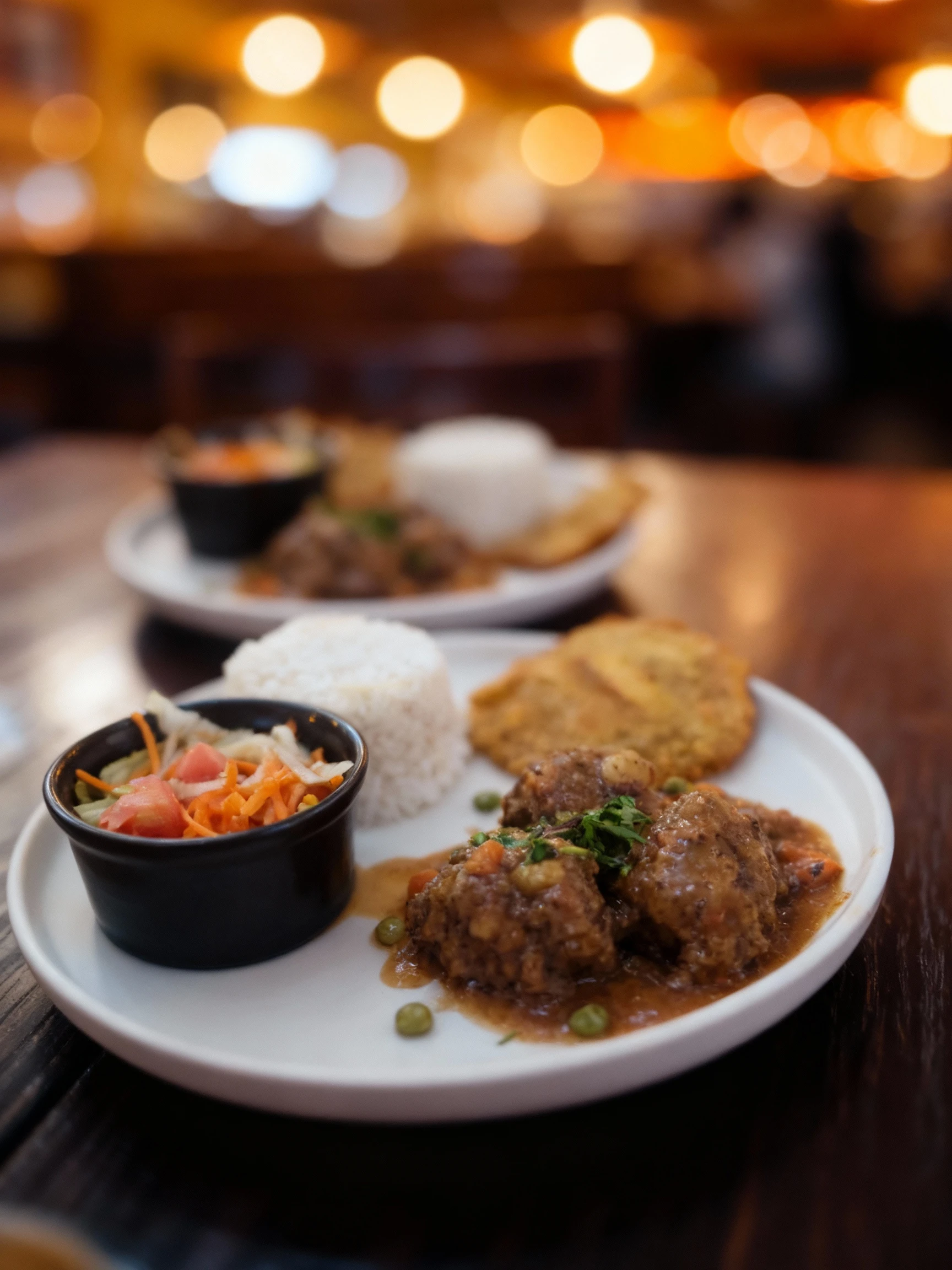 Two servings of a savory meat and vegetable stew (including carrots and peas) topped with fresh herbs, served with a mound of white rice, a fried plantain (tost