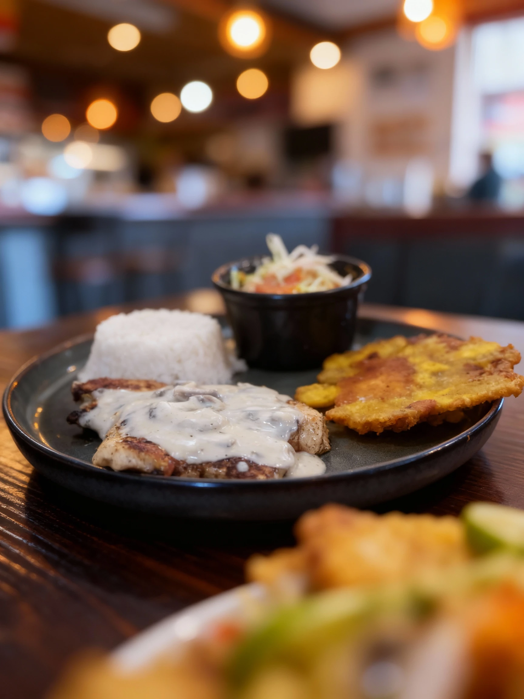 Grilled protein (likely chicken or steak) topped with a creamy mushroom sauce, served alongside a dome of white rice, a large fried plantain (tostone), and a si