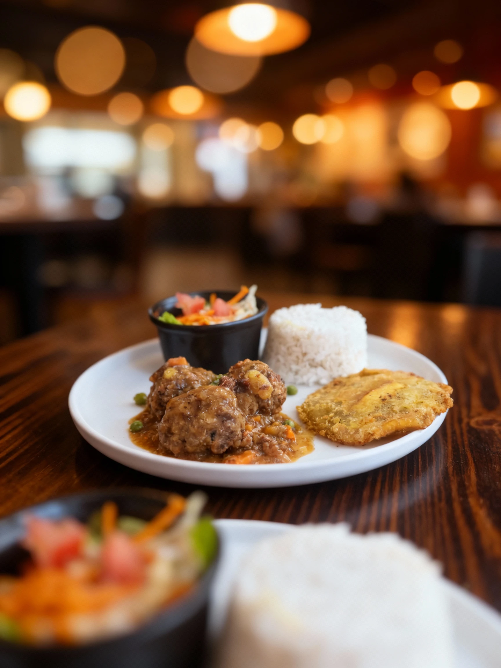 A plate featuring three meatballs stewed in a savory sauce with carrots and peas, accompanied by a molded portion of white rice and a large fried plantain (like