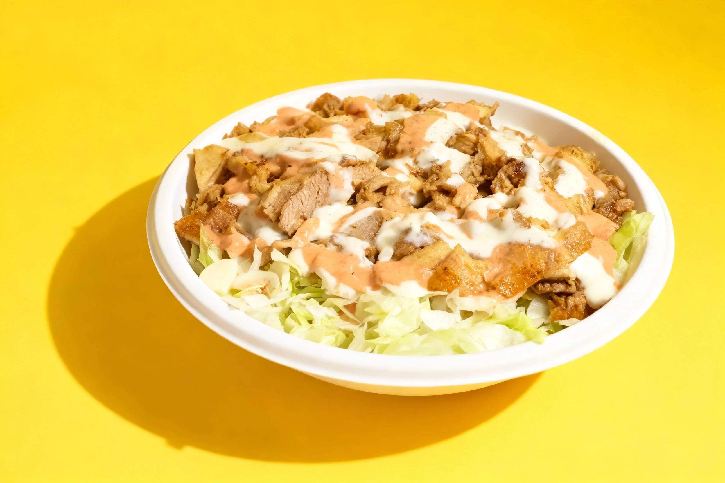 A gyro or shawarma plate featuring sliced, grilled meat served over shredded iceberg lettuce and heavily drizzled with white garlic sauce and a pinkish-orange s