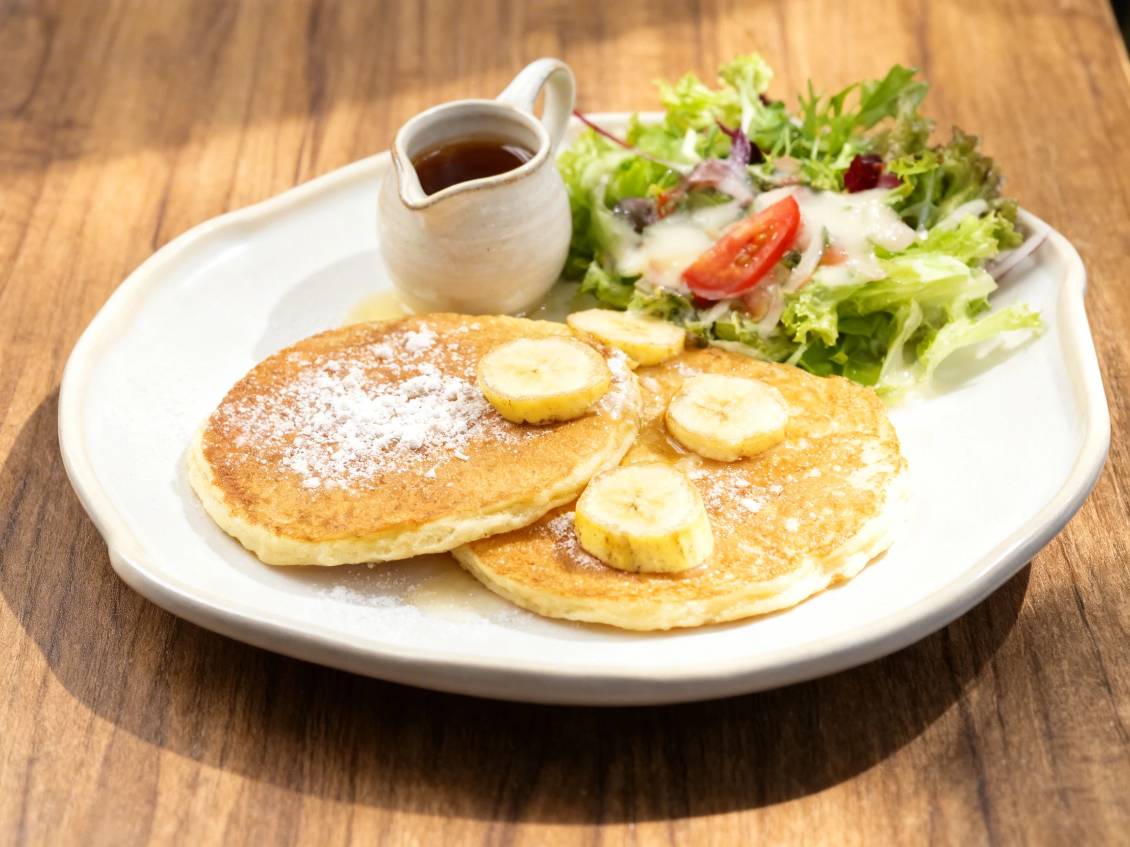 Fluffy pancakes topped with powdered sugar and sliced bananas, served with a side of syrup and a small garden salad.