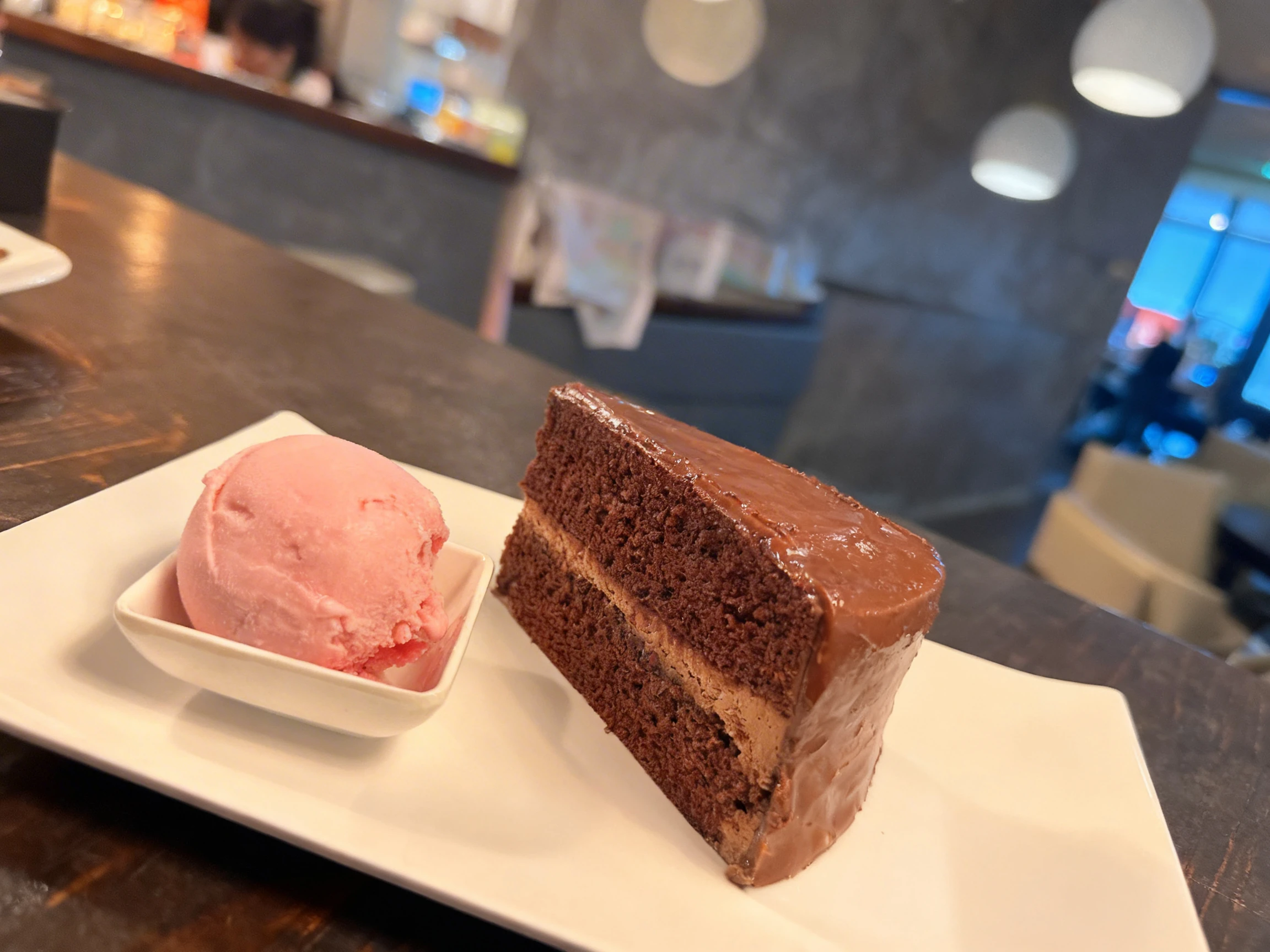 A slice of double-layer chocolate cake with chocolate frosting served alongside a scoop of pink strawberry sorbet in a small square dish.