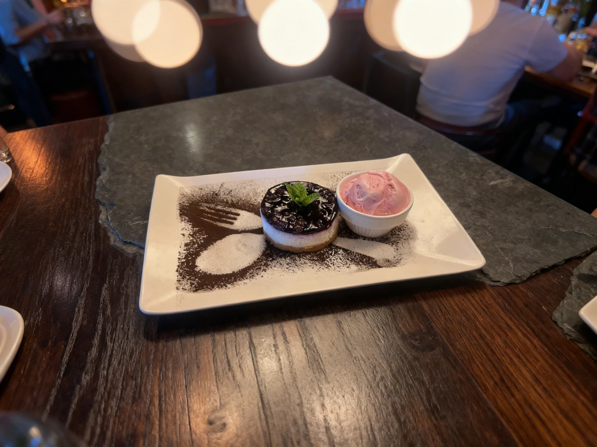 A round blueberry cheesecake topped with a mint sprig, served alongside a scoop of pink ice cream in a small ramekin. The white rectangular plate is decorated w