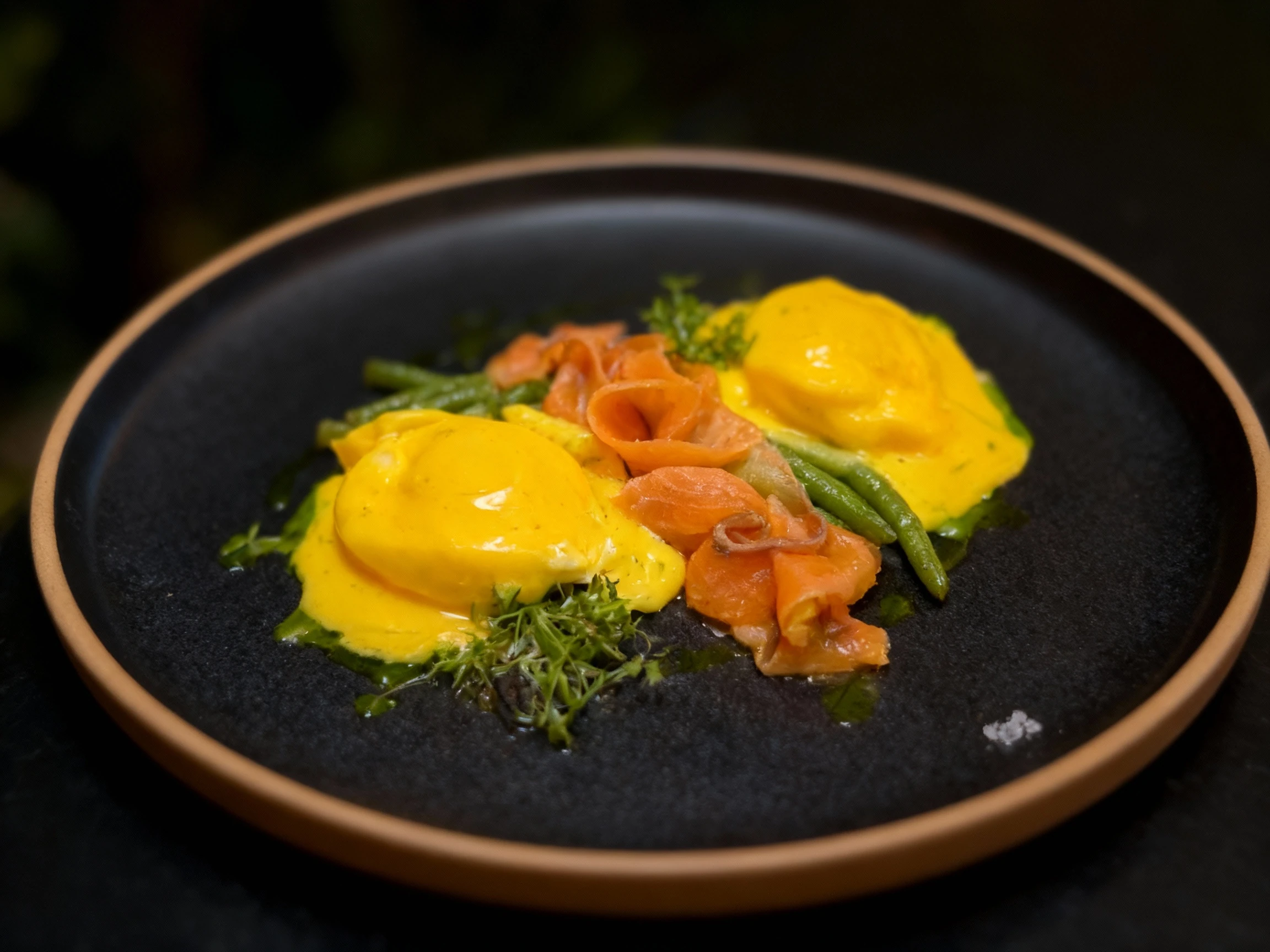 A refined dish featuring two poached eggs covered in rich Hollandaise sauce, topped with coiled smoked salmon and blanched green beans, garnished with fresh her