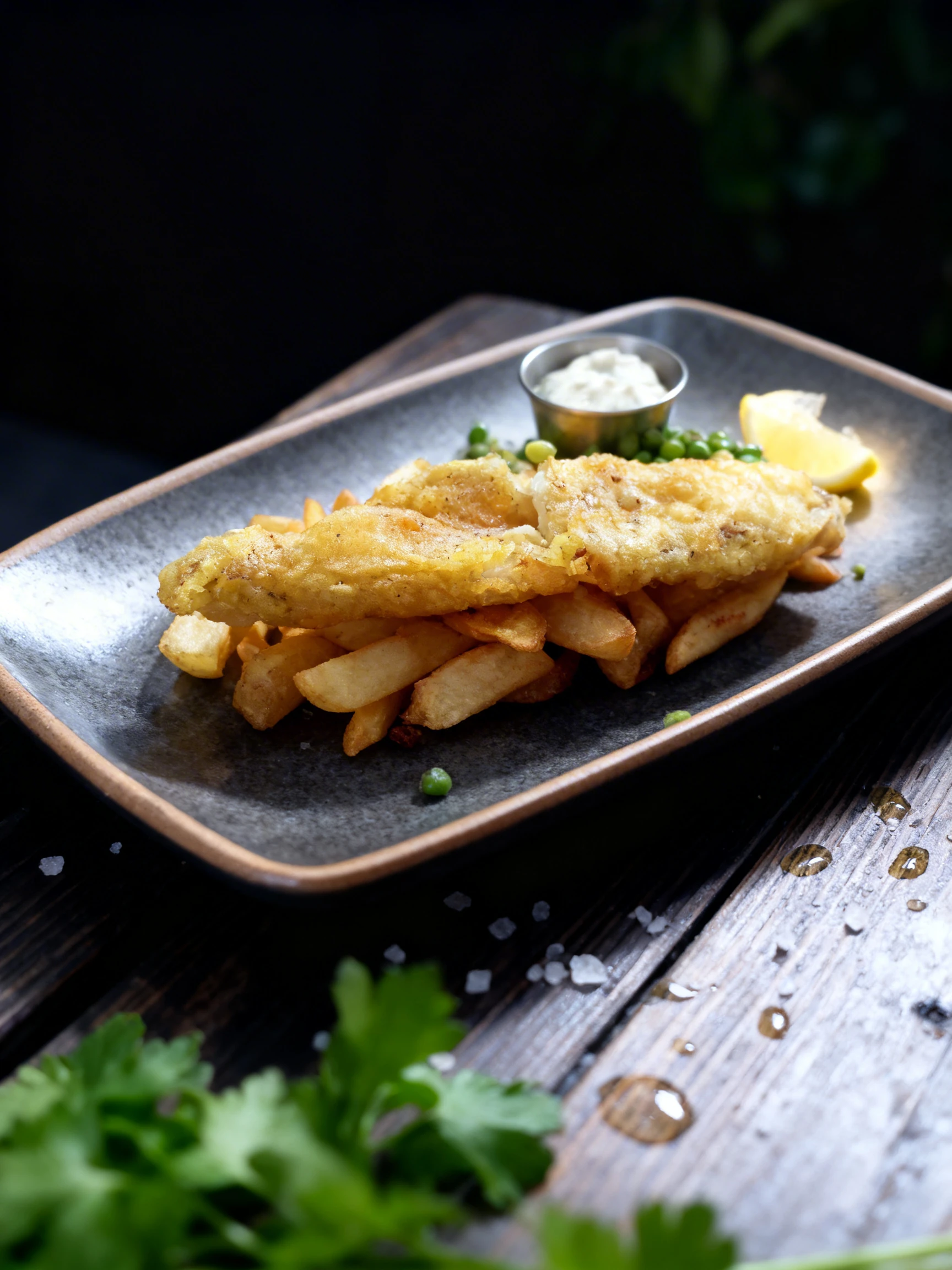 Battered fish fillets served over thick-cut chips with a side of green peas, tartare sauce, and a lemon wedge.