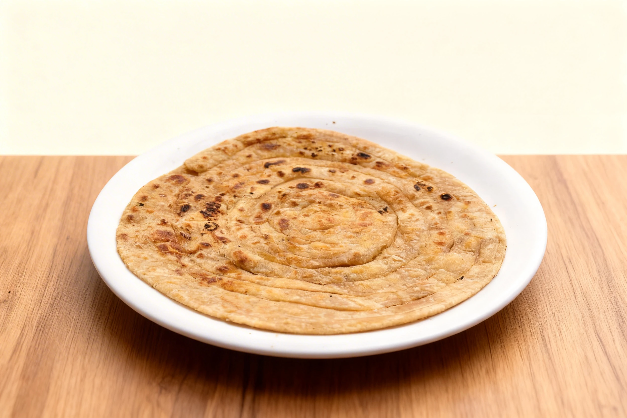 A circular, multi-layered Lachha Paratha with visible charred spots from being cooked on a tiddle or pan, served on a plain white plate.